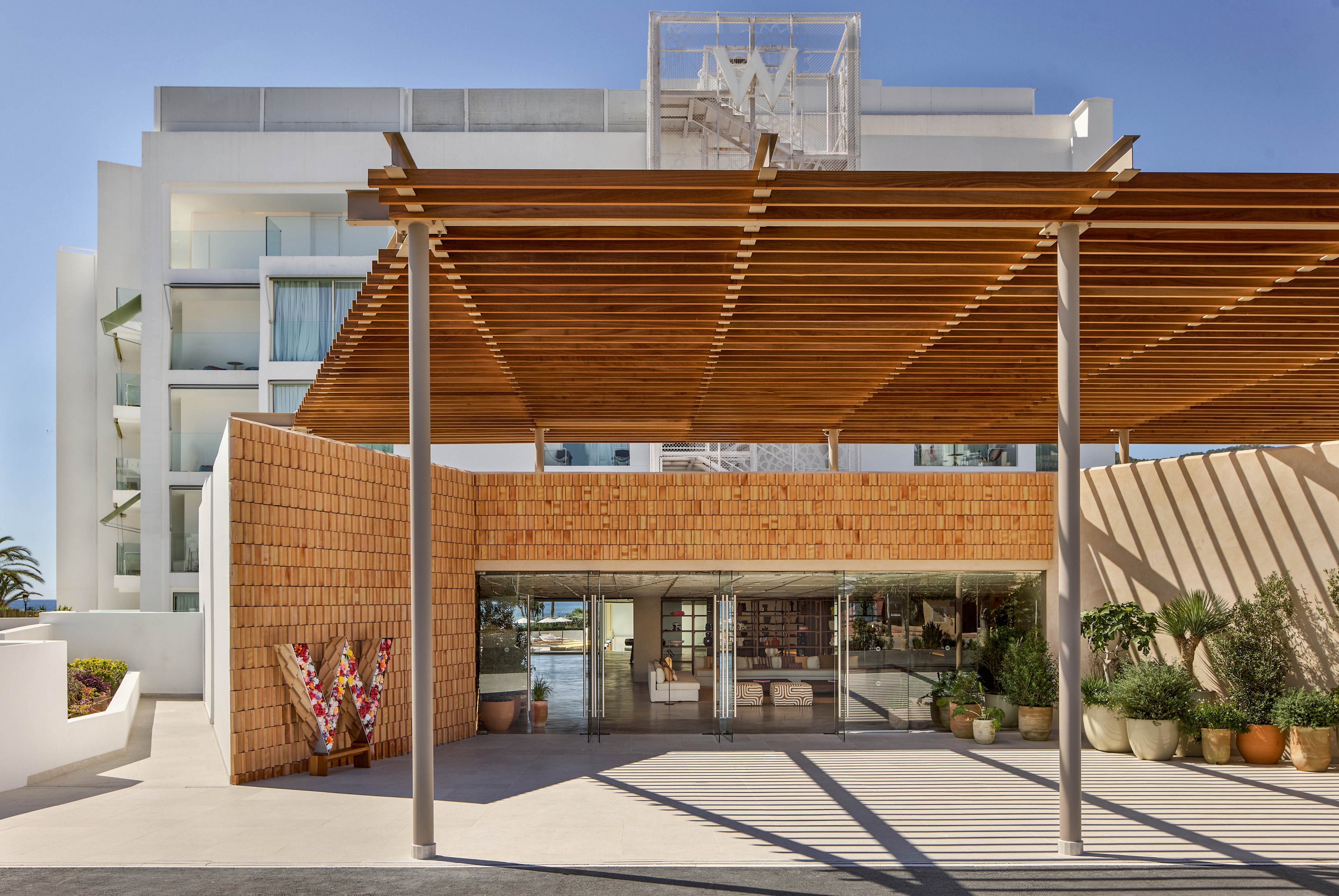 Contemporary hotel entrance featuring a wide wooden pergola leads into a glass-fronted lobby. The building facade combines white modern architecture with warm textured brick walls. Large potted plants line the walkway, adding greenery to the minimalist exterior. Bright daylight and clear skies create a clean, inviting resort atmosphere.