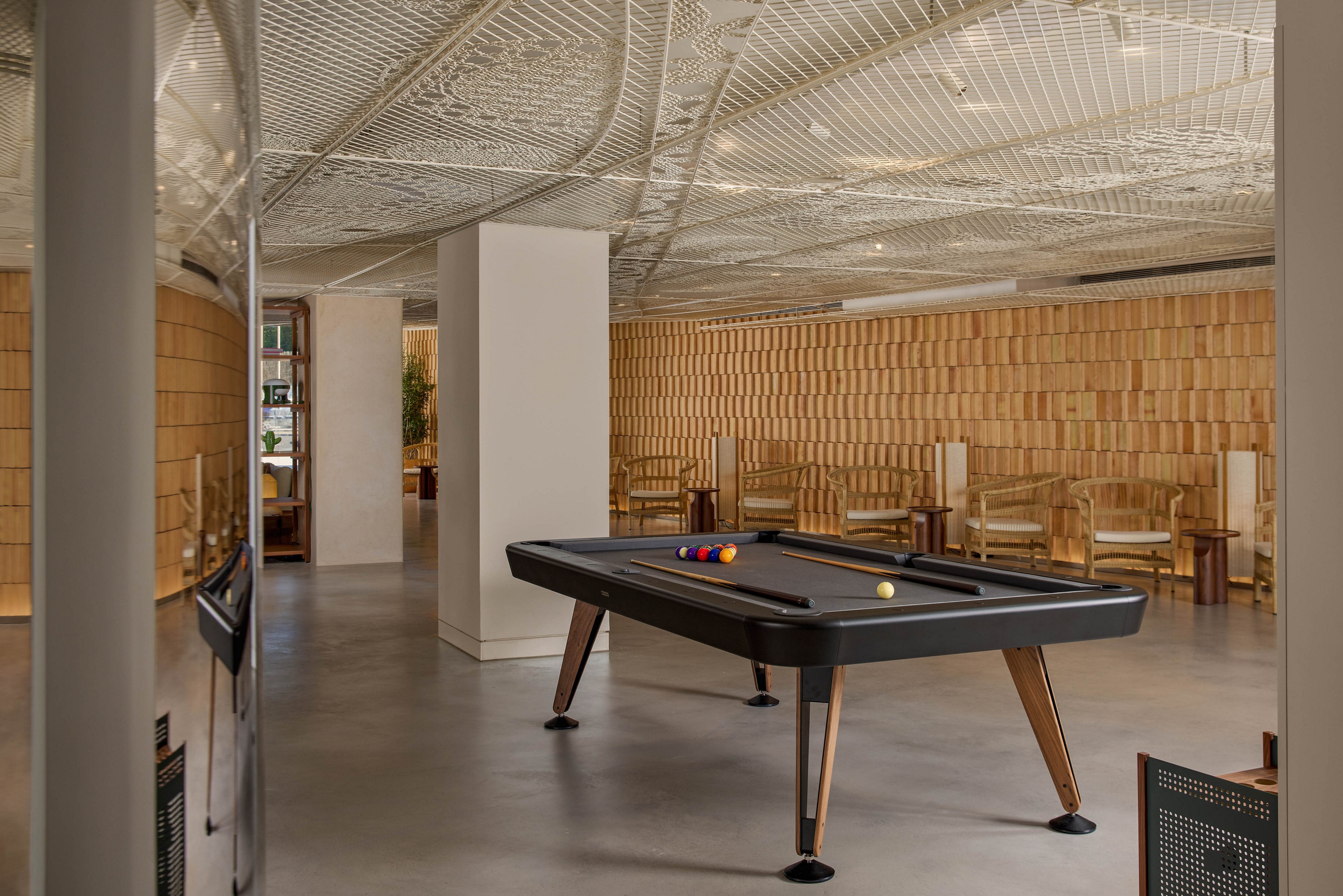 A contemporary lounge features a sleek black pool table as the central focus. The room has smooth concrete floors and warm wooden wall panels arranged in a geometric pattern. Minimalist wooden chairs line the walls, creating a relaxed social area. A sculptural, patterned ceiling adds visual interest and enhances the modern interior design.