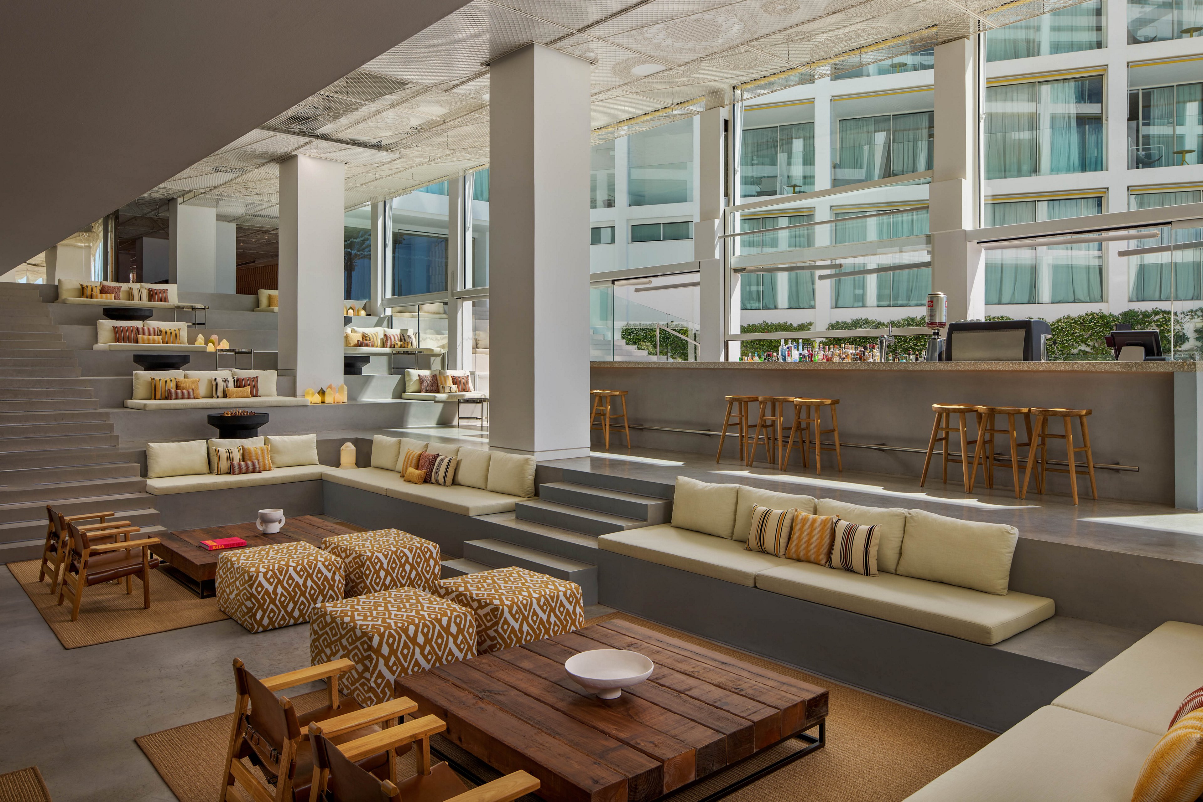 Contemporary lounge area features tiered seating with built-in sofas and cushioned platforms. A long bar with wooden stools lines one side of the space, creating a casual social atmosphere. Large floor-to-ceiling windows reveal an exterior building facade and bring in natural light. Neutral tones, patterned pillows, and wooden furniture contribute to a warm, modern aesthetic.