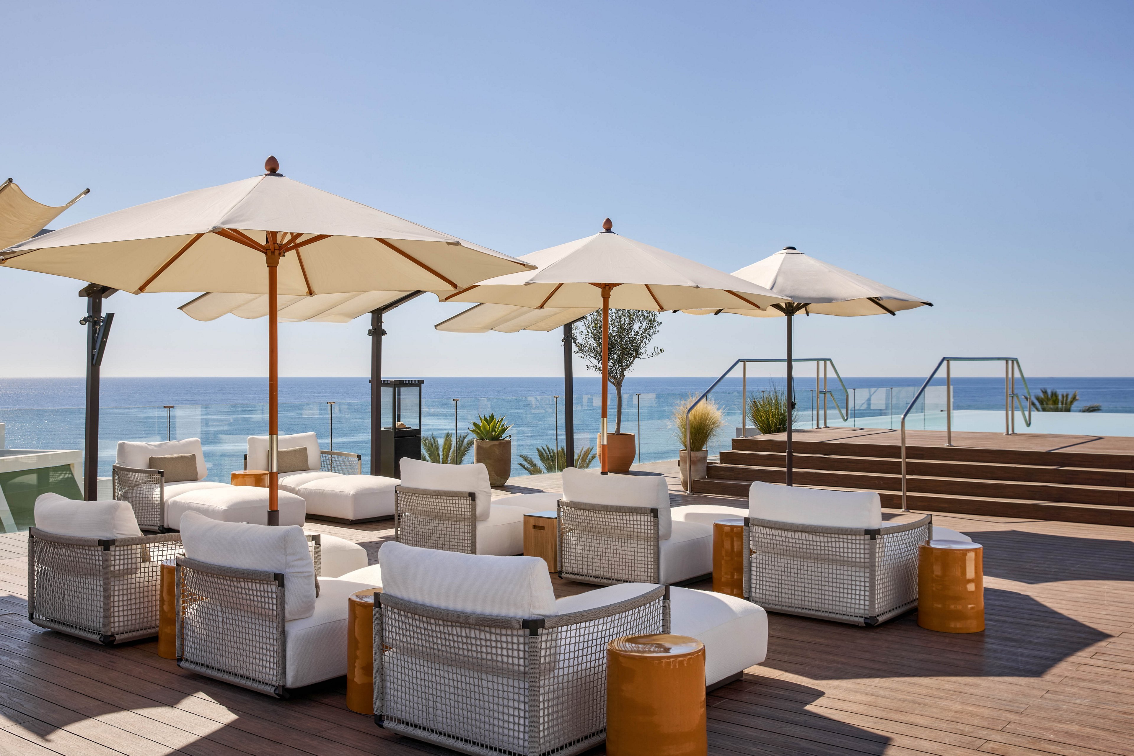 A stylish outdoor lounge area overlooks a calm blue sea under a clear sky. White cushioned chairs and wicker-style seating are arranged on a wooden deck around low side tables. Large white umbrellas provide shade, while steps lead up to a glass-railed platform with additional seating. The scene suggests a modern coastal resort or rooftop terrace designed for relaxation and ocean views.