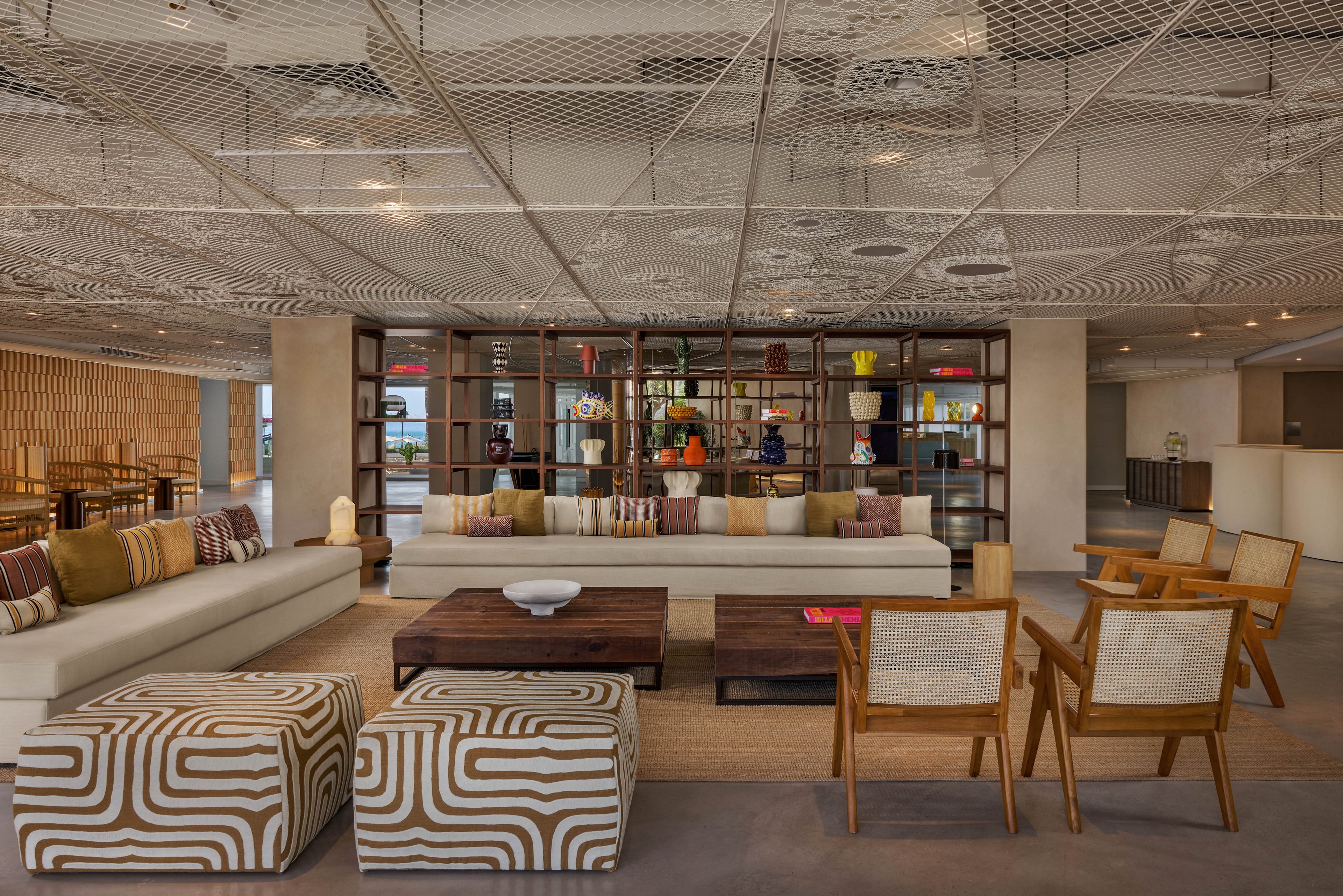 Spacious modern lobby features a long white sectional sofa accented with patterned cushions and low wooden tables. Two bold geometric ottomans and minimalist wooden armchairs create a relaxed lounge area in the foreground. A decorative open shelving unit with colorful vases and objects divides the space, while a mesh ceiling adds architectural interest. Warm neutral tones and soft lighting give the interior a calm, welcoming atmosphere.