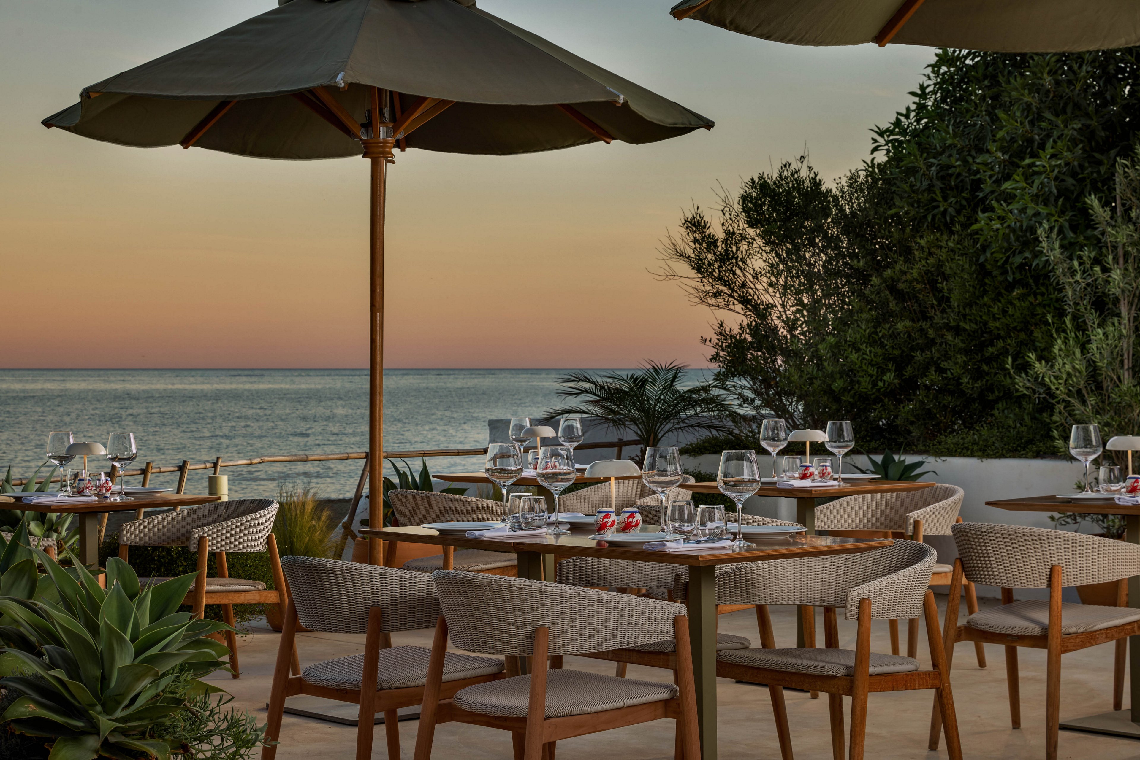 An upscale outdoor restaurant terrace overlooks a calm sea at sunset. Neatly arranged wooden tables and woven chairs sit beneath large patio umbrellas, each table set with wine glasses and tableware. Lush green plants frame the seating area, adding a natural touch to the sophisticated setting. The sky glows with soft pastel colors, creating a tranquil and romantic dining atmosphere by the water.