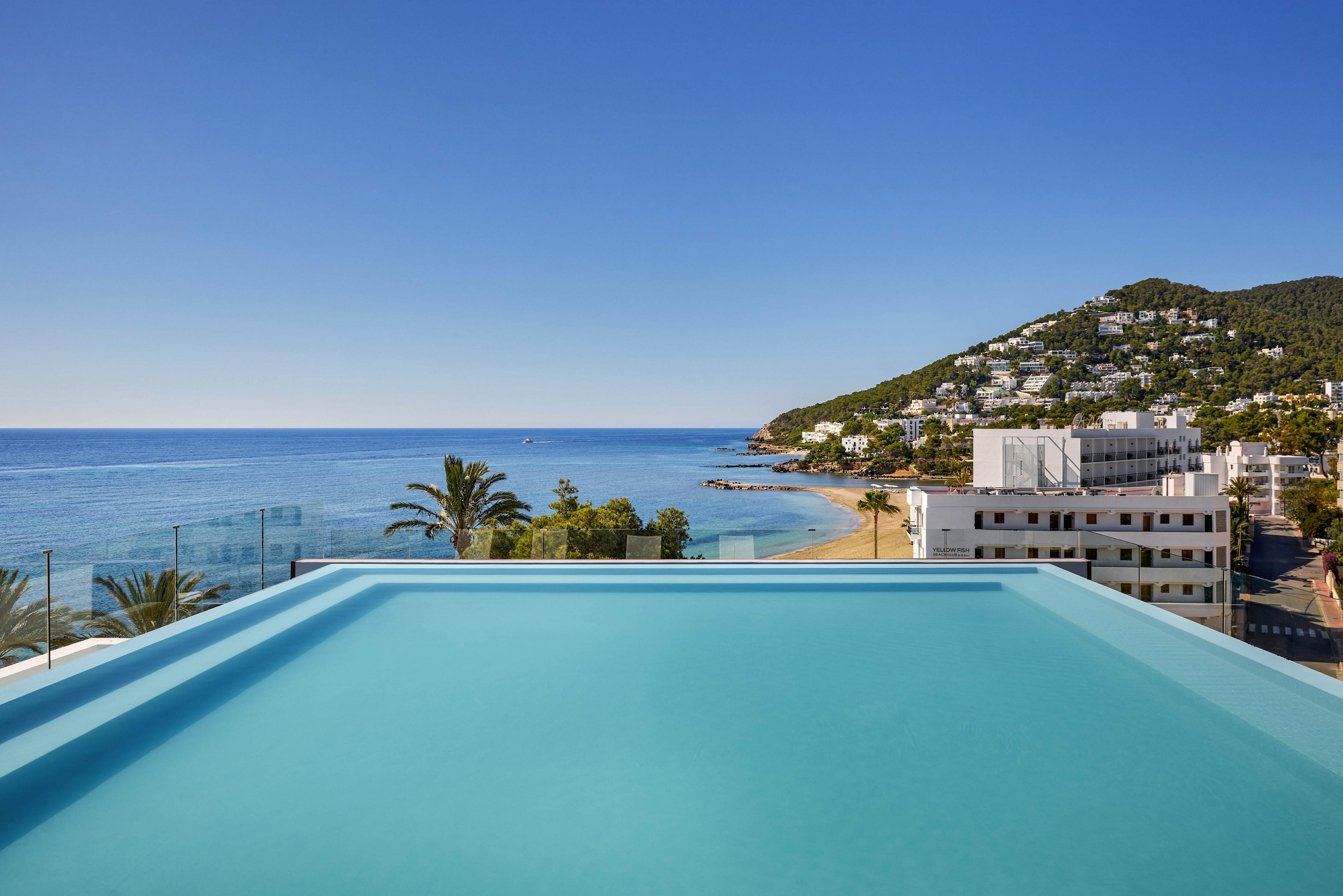 An infinity pool stretches toward a calm blue sea, creating a seamless horizon line. Beyond the pool, a sandy beach curves along the coastline beside low-rise white buildings. Hills covered with greenery and scattered houses rise in the background under a clear blue sky. The scene suggests a tranquil, modern seaside resort with a minimalist, sunlit atmosphere.