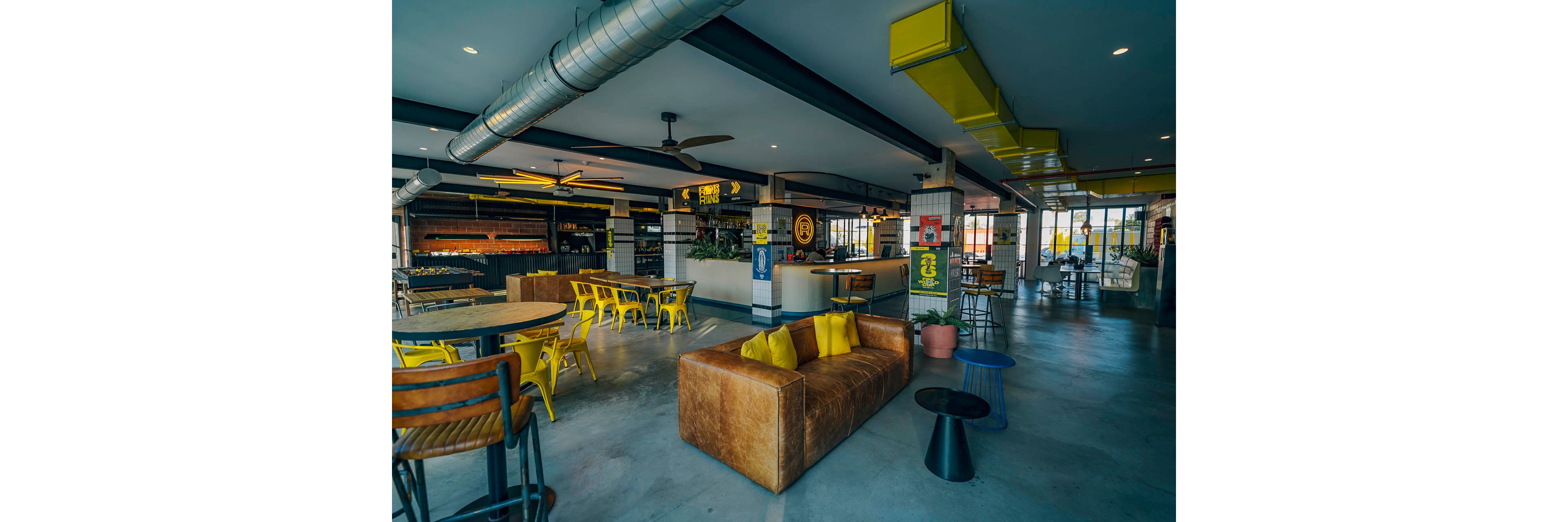 A contemporary restaurant interior features an industrial design with exposed ductwork and beams. A brown leather sofa with yellow pillows sits in the foreground beside small modern tables. In the background, wooden tables and bright yellow metal chairs create a casual dining area near the bar counter. Large windows bring in natural light, highlighting the concrete floor and colorful wall graphics.