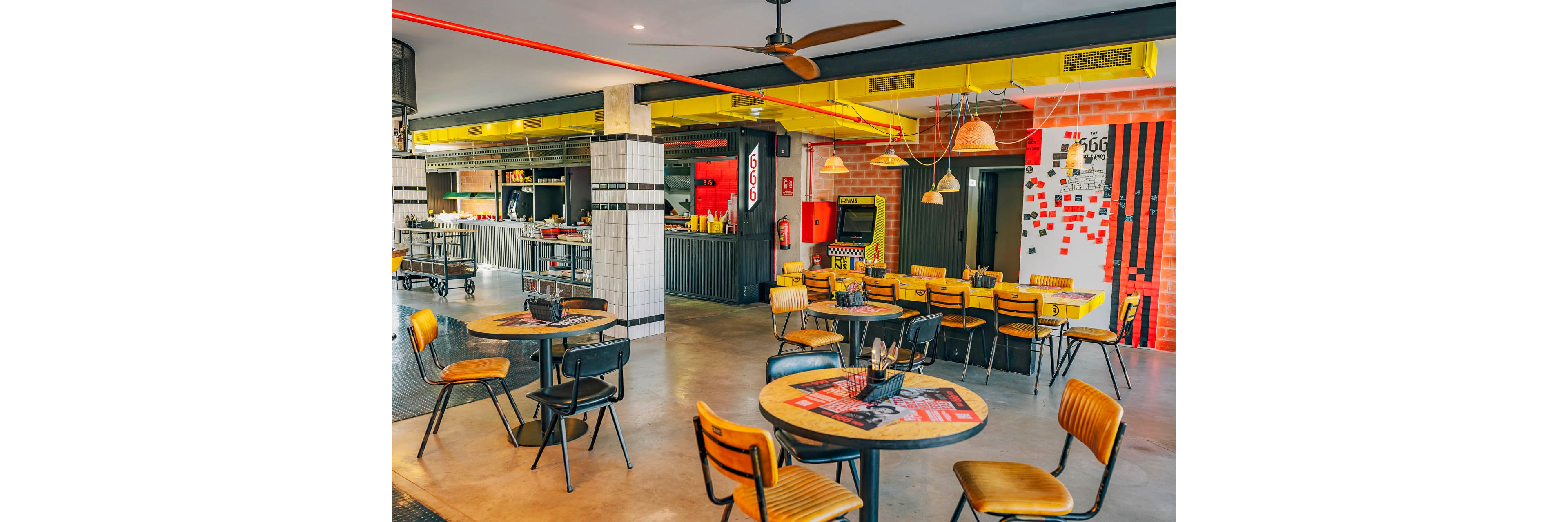 A vibrant cafeteria interior features several round wooden tables and metal chairs arranged in an open dining area. The setting includes an industrial-style counter and kitchen in the background, with tiled columns and exposed concrete elements. Bright yellow ceiling accents, red wall details, and hanging woven lamps create a lively, contemporary atmosphere. Menus and condiments are placed on the tables, while the space appears clean, organized, and ready for customers.