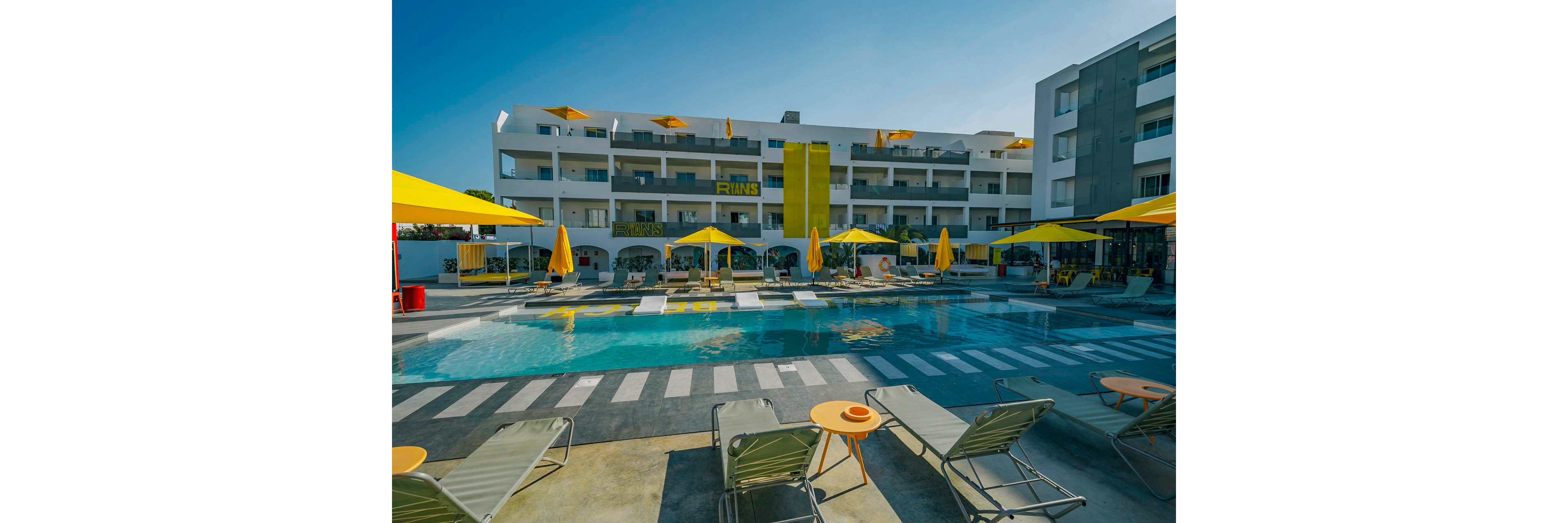 A contemporary hotel courtyard features a central swimming pool surrounded by lounge chairs and bright yellow sun umbrellas. The multi-story white building in the background has balconies and bold yellow accents, including a large vertical stripe. Clear blue sky and strong sunlight create a vibrant, summery atmosphere. The scene suggests a relaxed vacation setting with ample seating and shaded areas for guests.
