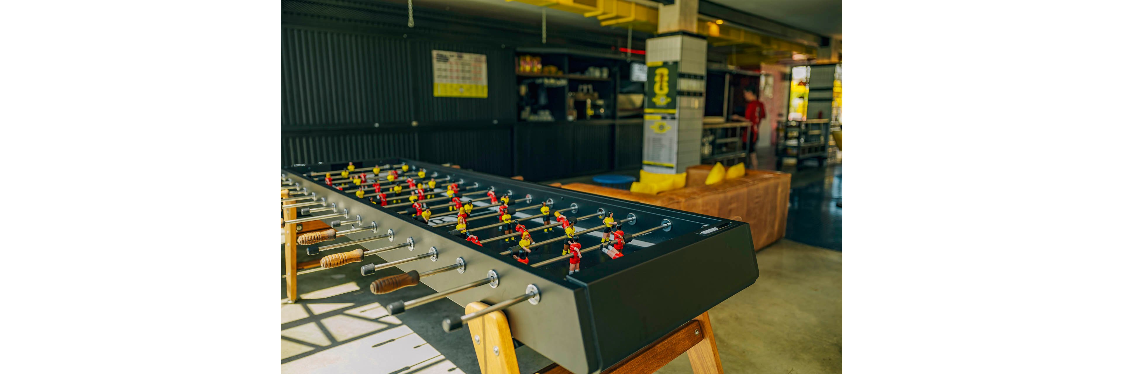 A large foosball table with wooden legs stands prominently in a bright, modern lounge area. Small red and yellow player figures line the metal rods across the dark tabletop. In the background, casual seating with cushions and industrial-style decor create a relaxed social atmosphere. Sunlight streams in from the side, adding warm tones to the contemporary recreational space.