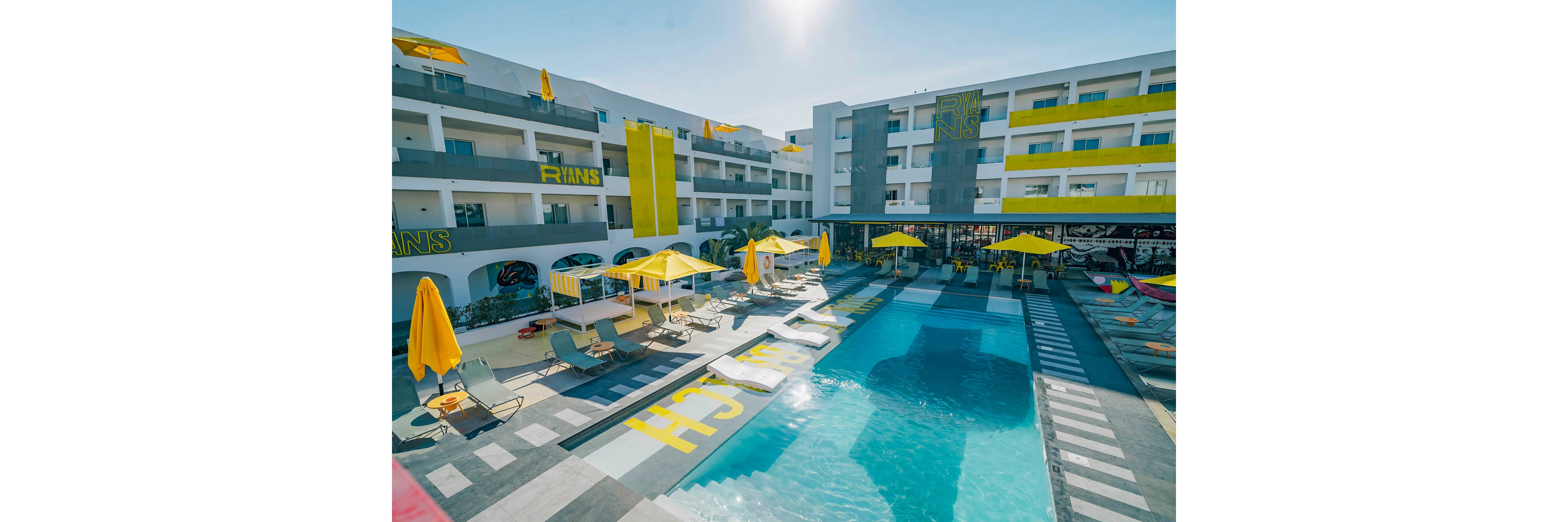A contemporary hotel courtyard features a central swimming pool surrounded by lounge chairs and yellow umbrellas. The multi-story white building has balconies overlooking the pool and bold yellow accents on walls and awnings. Visible text such as "RIANS" and large yellow letters on the ground add a graphic, playful touch. The bright daylight and clear blue sky create a vibrant, summery atmosphere ideal for vacation relaxation.