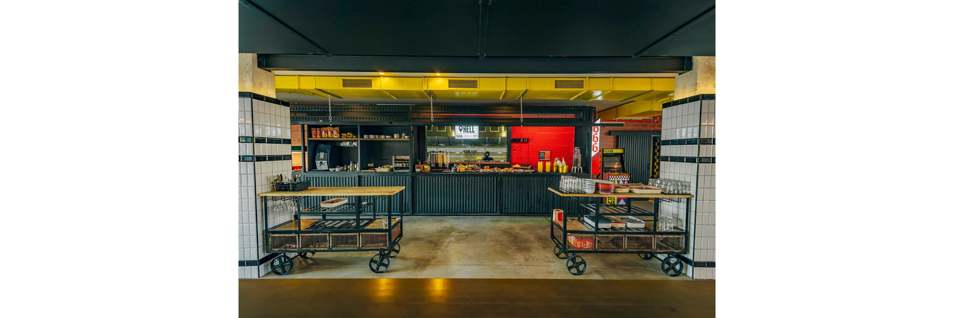 A modern cafeteria counter is set in an industrial-style interior with corrugated metal panels and exposed ceiling ducts. Two wheeled service carts stand in the foreground, holding trays, glasses, and condiments. The back counter displays various food items, drink dispensers, and shelves with snacks. Bright yellow and red accent colors contrast with the dark metal and white tiled columns, creating a contemporary urban atmosphere.