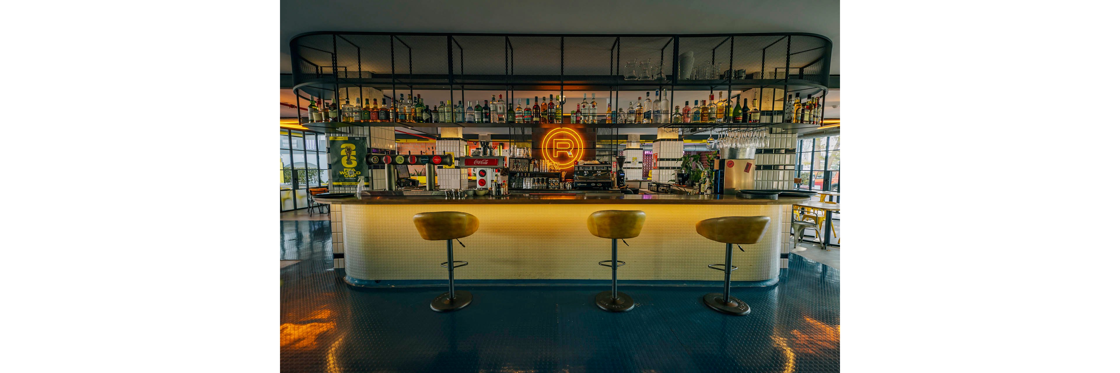A contemporary bar interior features a long counter with three mustard-yellow bar stools in front. Shelves behind the bar are filled with assorted liquor bottles and glassware, framed by a black metal structure. A bright circular neon sign with a large letter R glows at the center of the back bar, adding a focal point. The space has a polished blue floor and warm ambient lighting, creating a stylish, inviting atmosphere.
