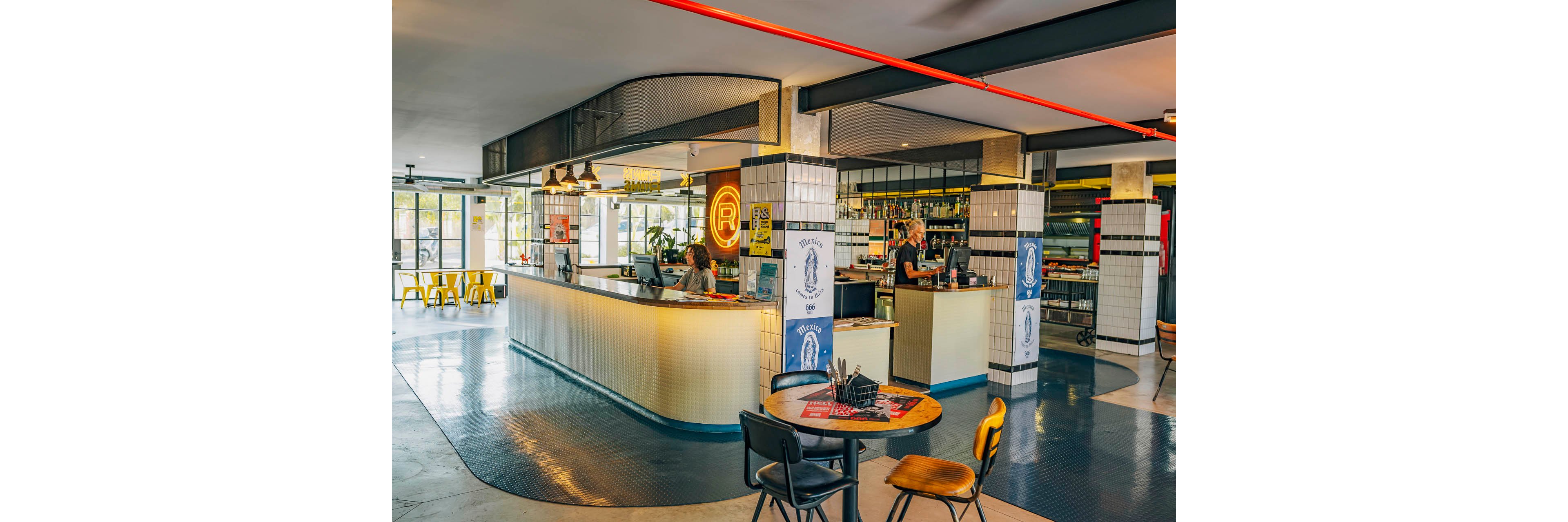 A contemporary bar counter dominates the interior of a bright, open venue. Bartenders work behind the tiled bar, with shelves of bottles and glassware visible in the background. A round table with chairs sits in the foreground, and large windows bring in natural light from outside. Warm neon lighting, clean lines, and colorful accents create a relaxed, modern atmosphere.