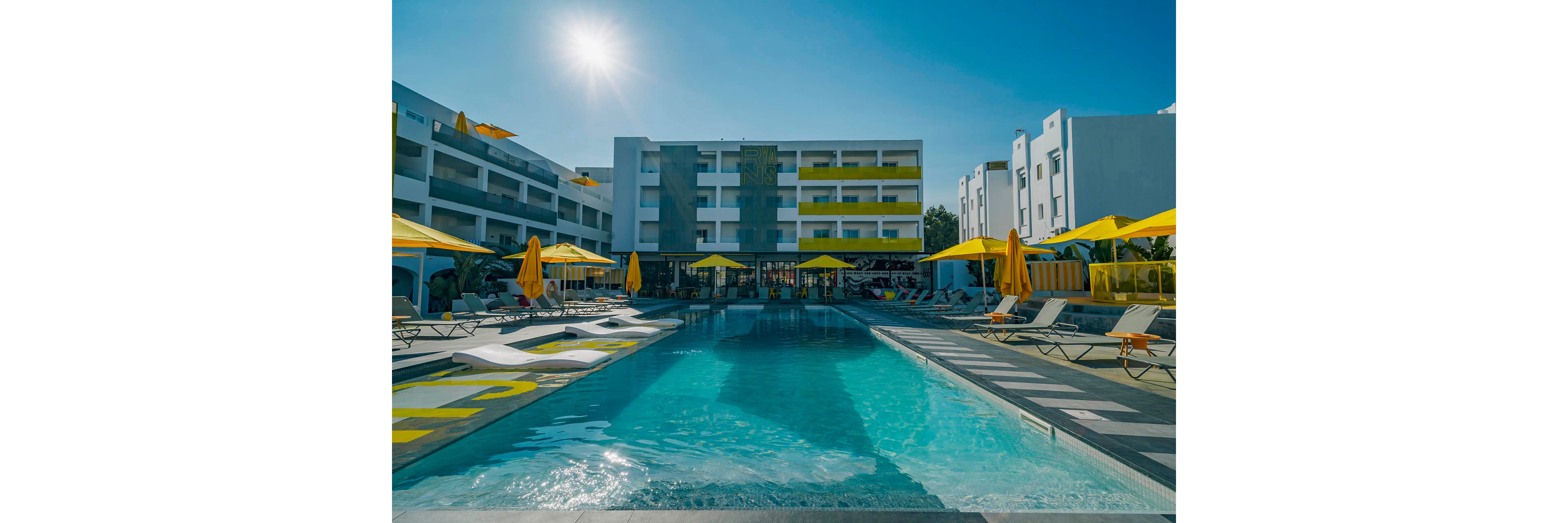 A contemporary hotel courtyard features a central swimming pool surrounded by sun loungers and bright yellow umbrellas. The multi-story white and gray building with yellow accents frames the pool area, creating a clean geometric look. Clear blue water reflects the sunny sky, suggesting a warm, relaxing vacation atmosphere. The scene is calm and inviting, with no people visible around the poolside.
