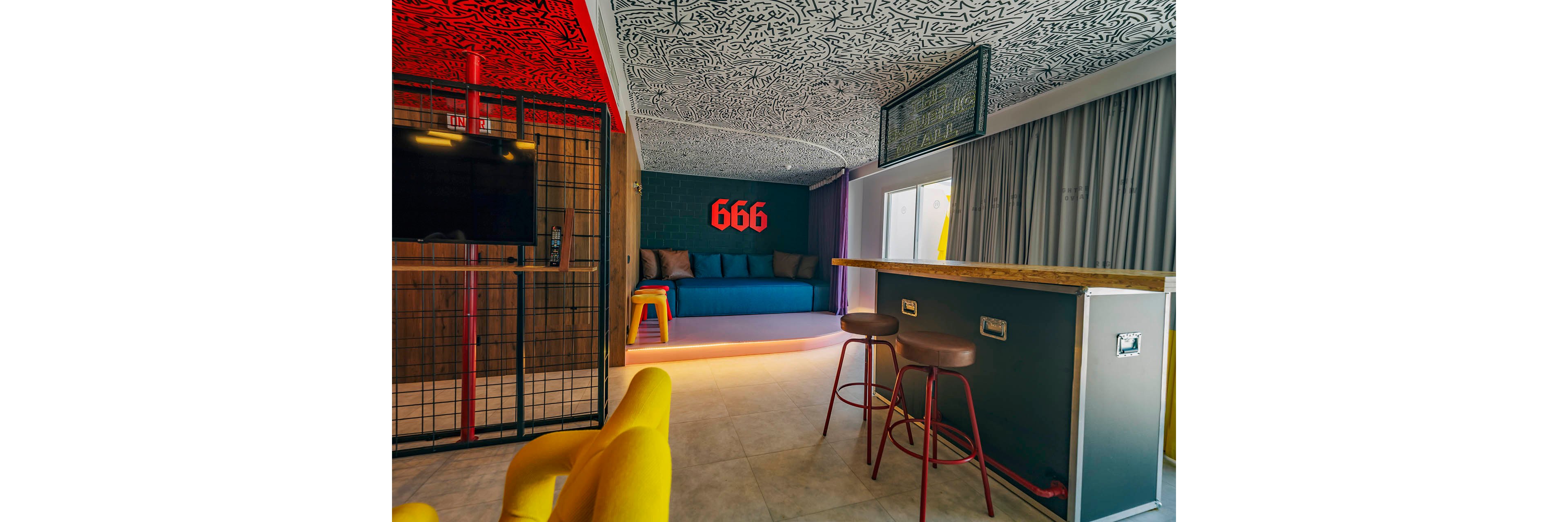 A contemporary lounge features a blue sofa against a dark accent wall with the red number 666 prominently displayed. In the foreground, a high counter with red bar stools faces a wall-mounted television enclosed in a metal grid. Bright yellow chairs and a yellow side stool add bold color contrast to the space. The ceiling is decorated with intricate black-and-white patterns, while large windows and curtains bring in natural light, creating a vibrant, eclectic interior.