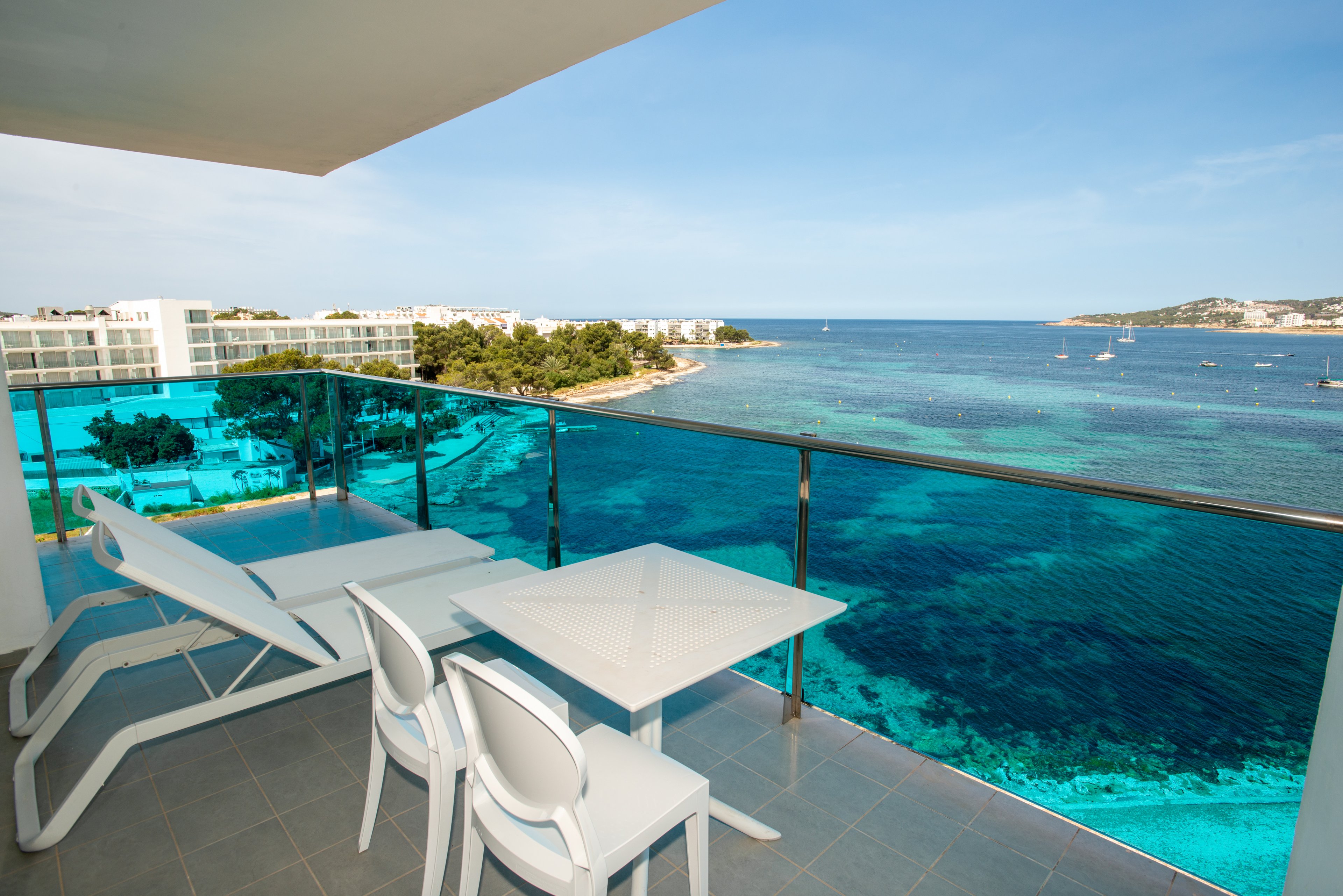 A modern balcony overlooks a clear turquoise sea and distant shoreline. Two white lounge chairs and a small table with chairs are arranged on the tiled terrace, framed by a glass railing. In the background, low-rise resort buildings and trees line the coast under a bright blue sky. Several small boats are visible on the calm water, enhancing the relaxed vacation atmosphere.