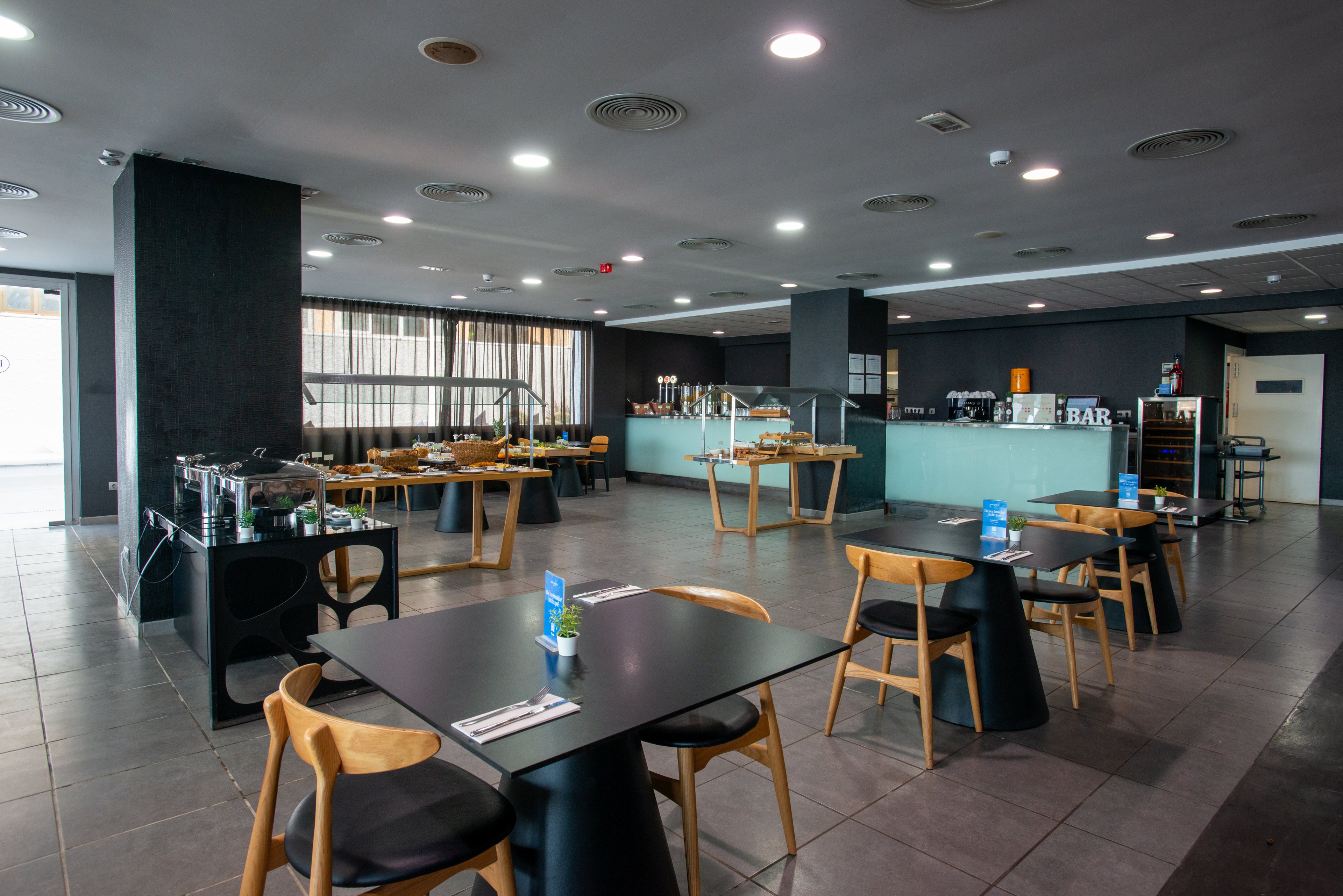 Spacious contemporary restaurant interior with neatly arranged wooden tables and chairs. The room appears to be part of a hotel dining or breakfast area, with buffet stations set up along the walls. A bar counter is visible in the background, along with coffee machines and drink dispensers. The space is brightly lit with large windows, tiled flooring, and a clean, minimalist design.