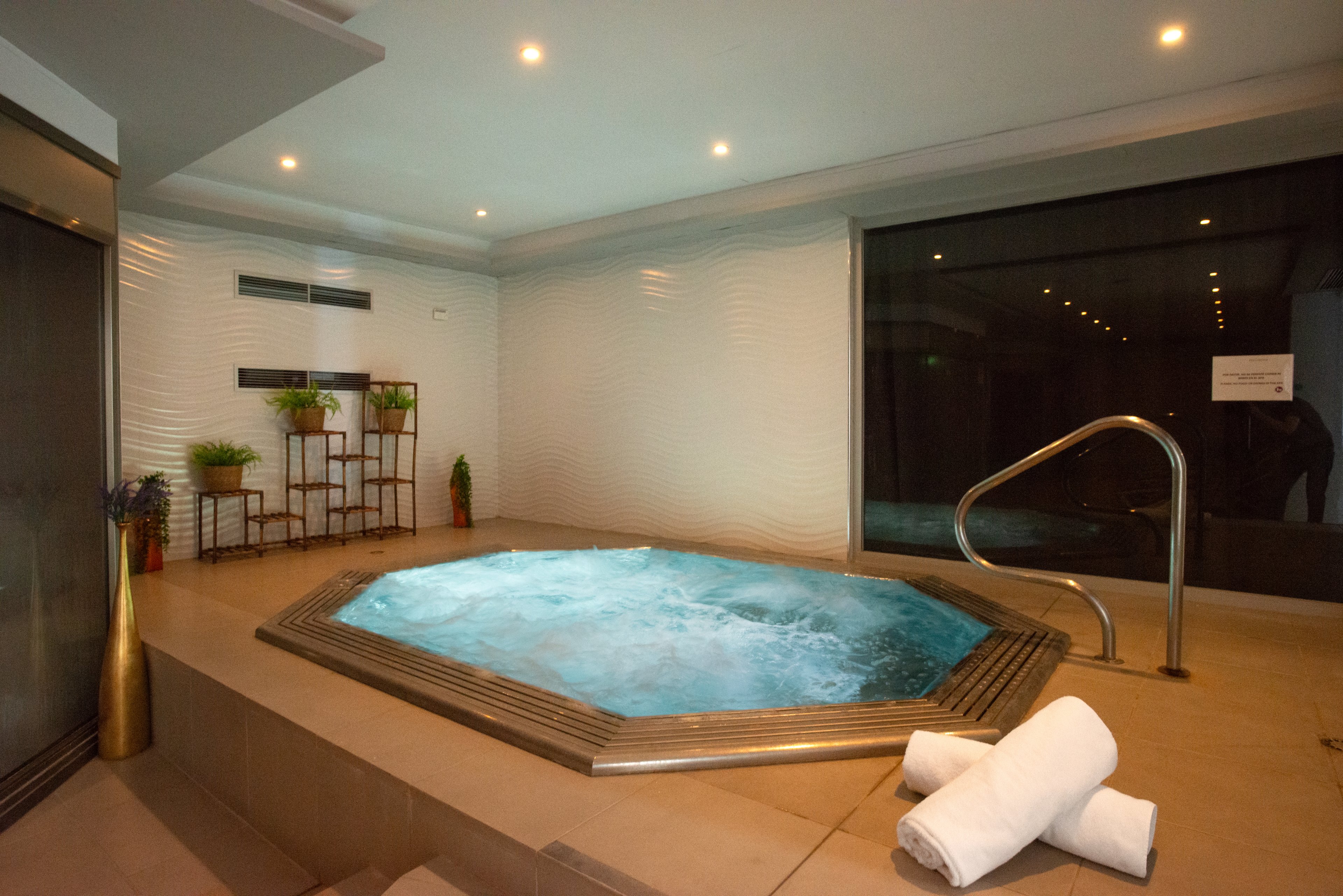 An indoor spa room features a bubbling jacuzzi set into a tiled platform. Rolled white towels rest on the edge of the hot tub, ready for guests. Soft recessed ceiling lights and a large dark window create a calm, intimate atmosphere. Decorative plants and modern fixtures enhance the relaxing, contemporary spa setting.