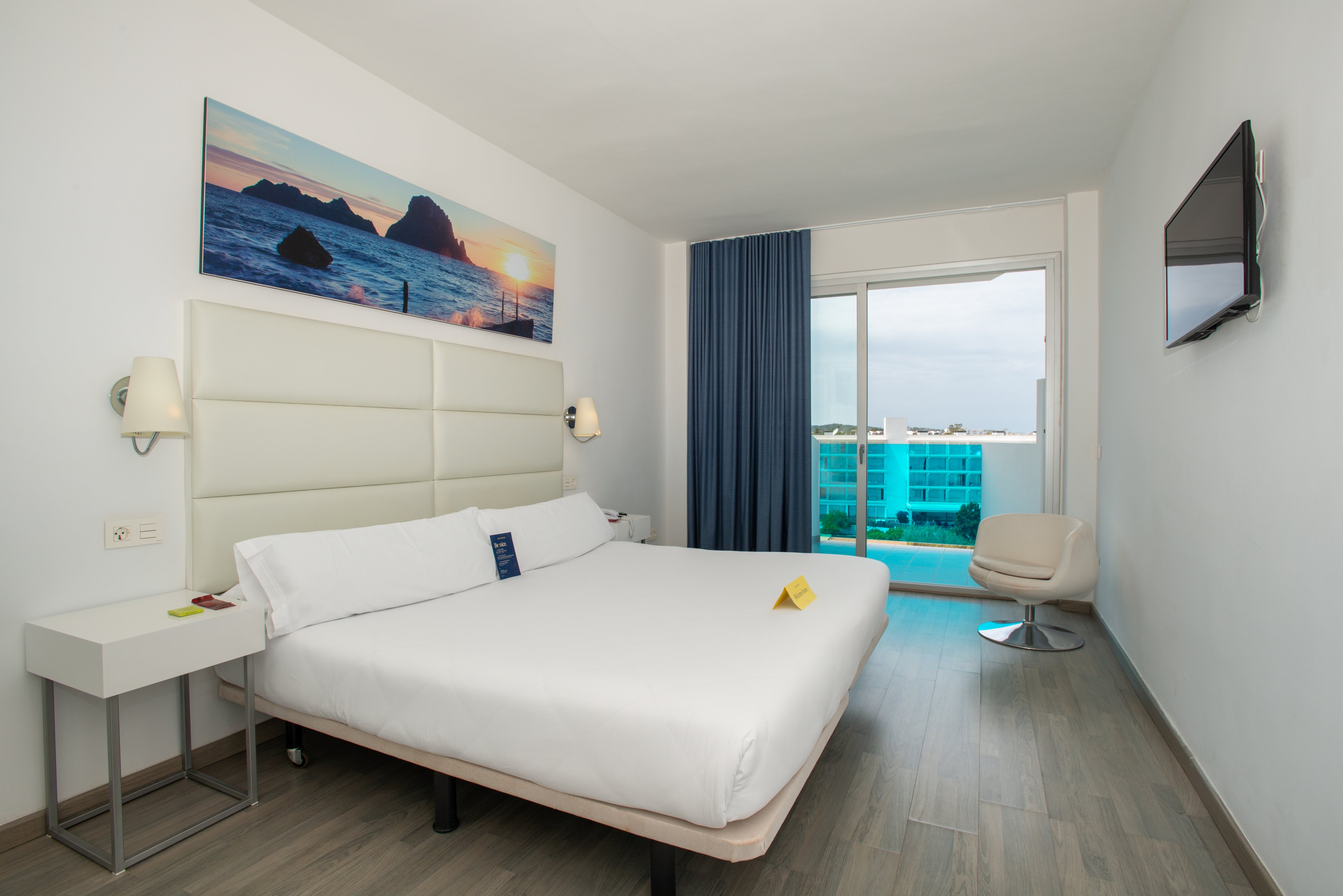 Bright contemporary hotel room featuring a large double bed with white linens and a padded headboard. The space includes a bedside table, wall-mounted lamps, and a flat-screen TV on a white wall. A sliding glass door opens to a balcony overlooking a pool and modern buildings outside. Blue curtains, wood-style flooring, and a seascape artwork above the bed create a clean, relaxing atmosphere.