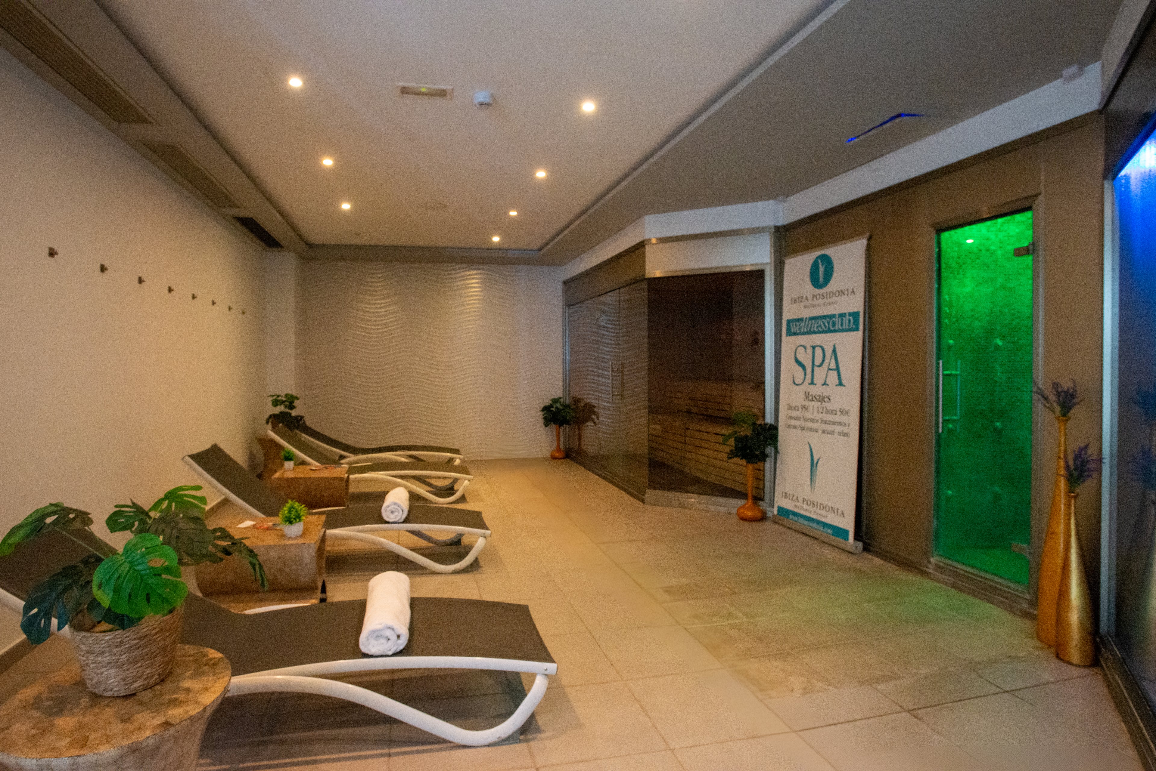 Indoor spa relaxation area featuring several reclining lounge chairs with rolled white towels. The space includes potted plants, soft ceiling lighting, and a tiled floor creating a clean, modern look. A glass-enclosed sauna or steam room and a green-lit shower or treatment room are visible in the background. A vertical banner with the word SPA and additional text stands near the treatment rooms, emphasizing the wellness setting.