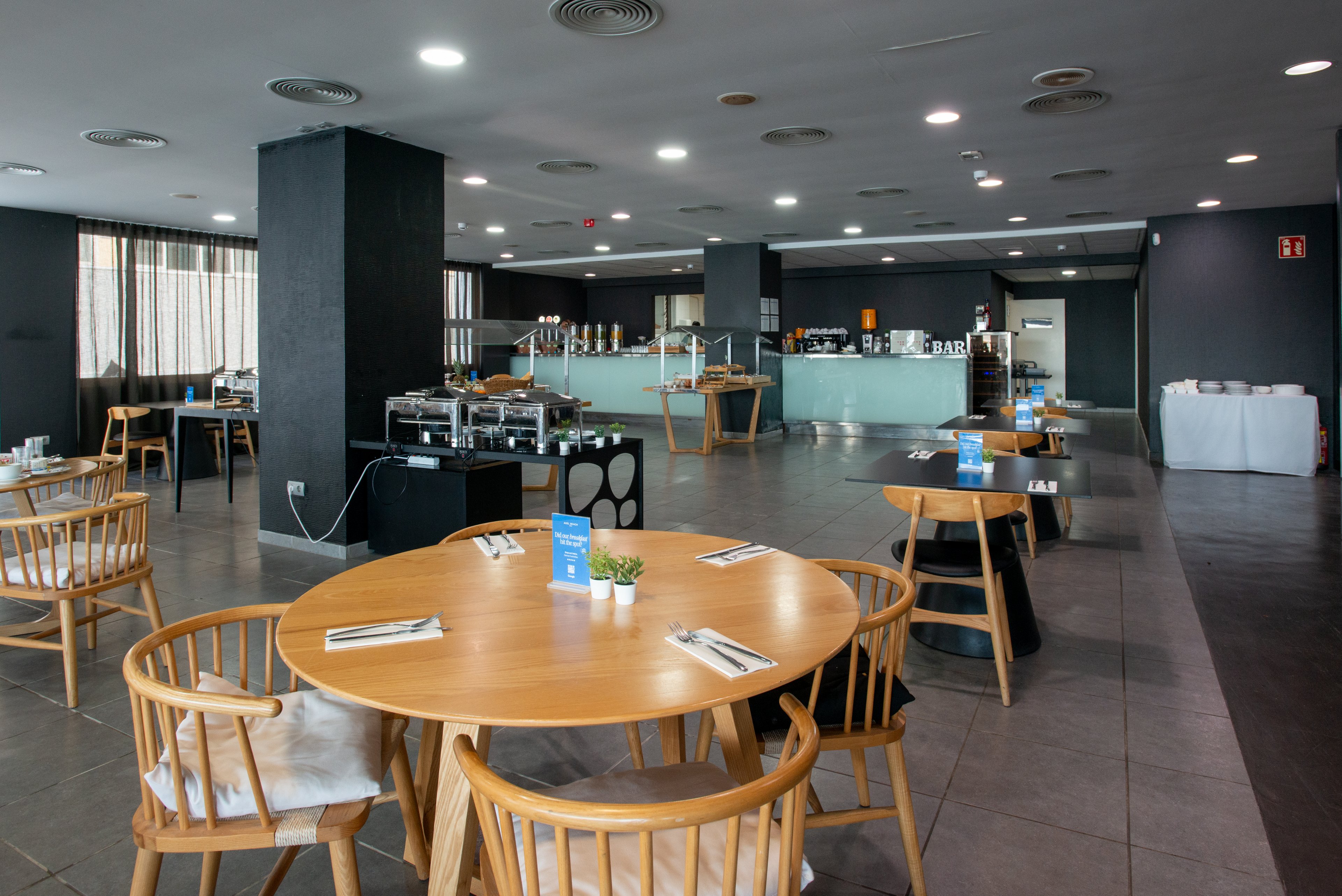 Spacious contemporary restaurant interior featuring round wooden tables and chairs. The setting appears to be a hotel dining room with a self-service buffet counter in the background. Neutral gray walls and flooring contrast with light wood furniture for a clean, minimalist look. Table settings with cutlery, napkins, and small potted plants add a casual yet organized atmosphere.