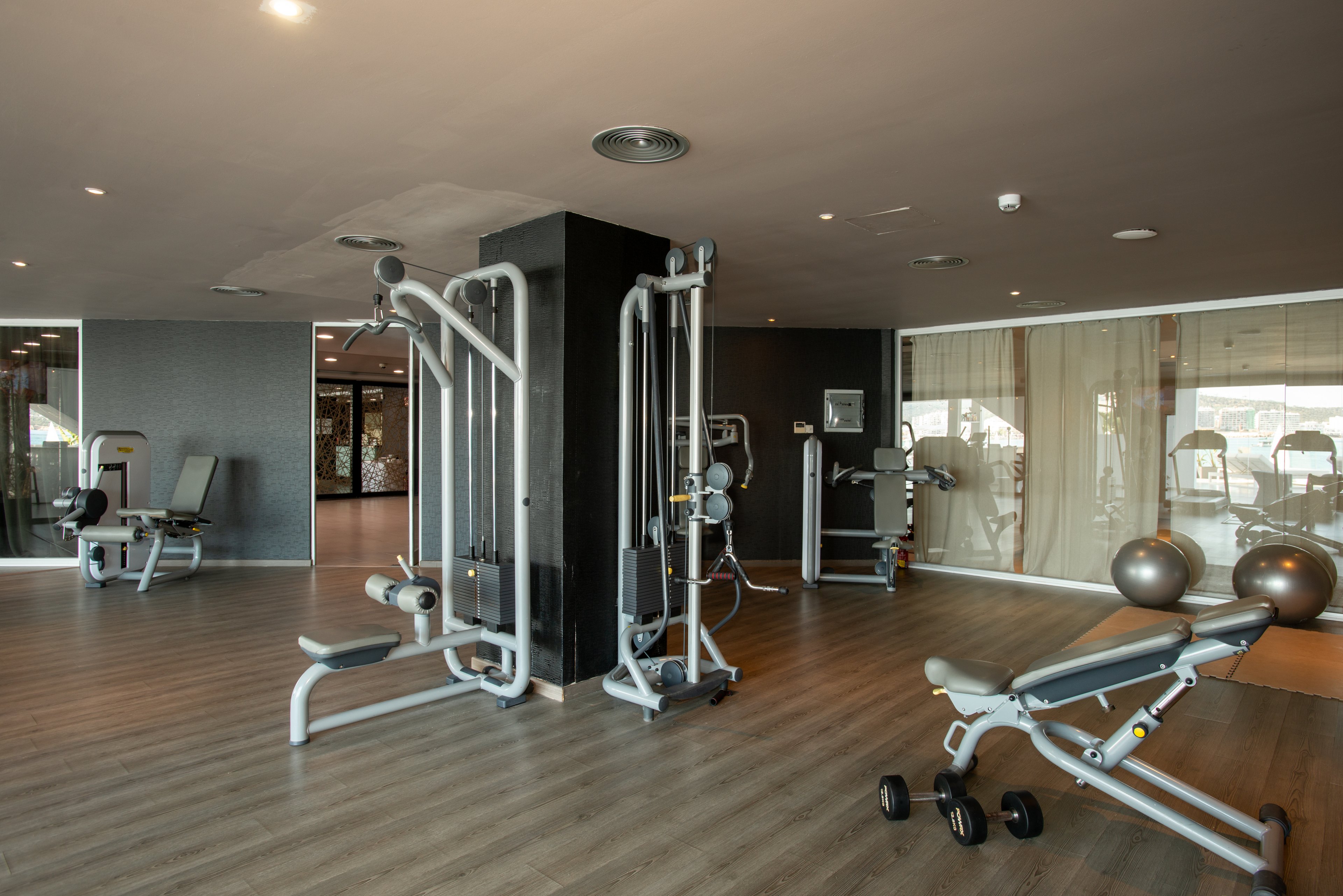 A spacious, modern gym interior features several strength-training machines arranged around a central column. The room has wood-style flooring and neutral-colored walls, creating a clean and minimalist atmosphere. Large windows in the background allow natural light to illuminate treadmills and other cardio equipment. Two exercise balls and a bench add variety to the workout options in this quiet, empty fitness space.