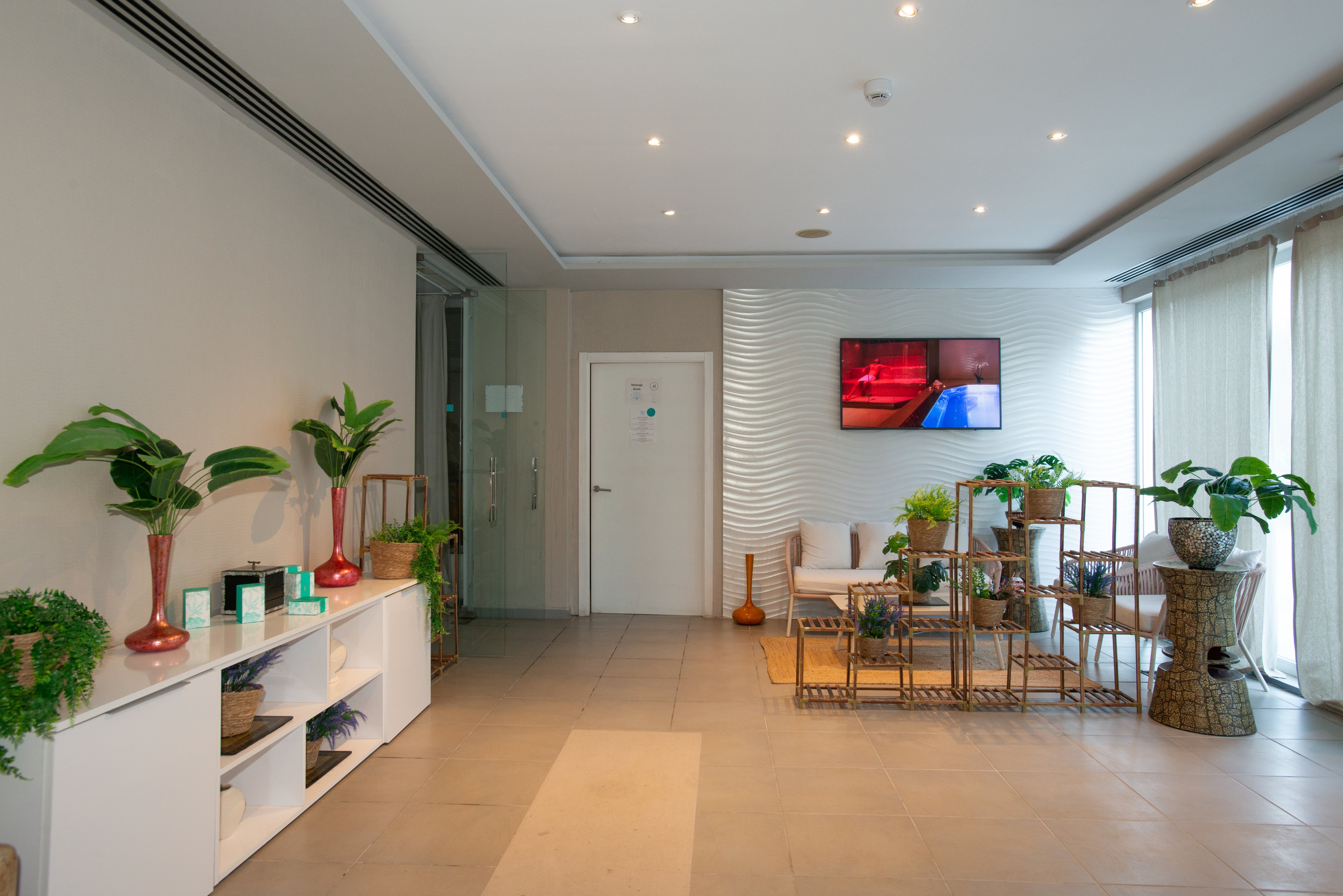 A contemporary spa waiting area features comfortable seating and abundant indoor plants. Neutral walls and tiled flooring create a calm, minimalist atmosphere. A textured white accent wall and a wall-mounted television add visual interest to the space. Natural light filters through sheer curtains, enhancing the relaxing, wellness-focused environment.