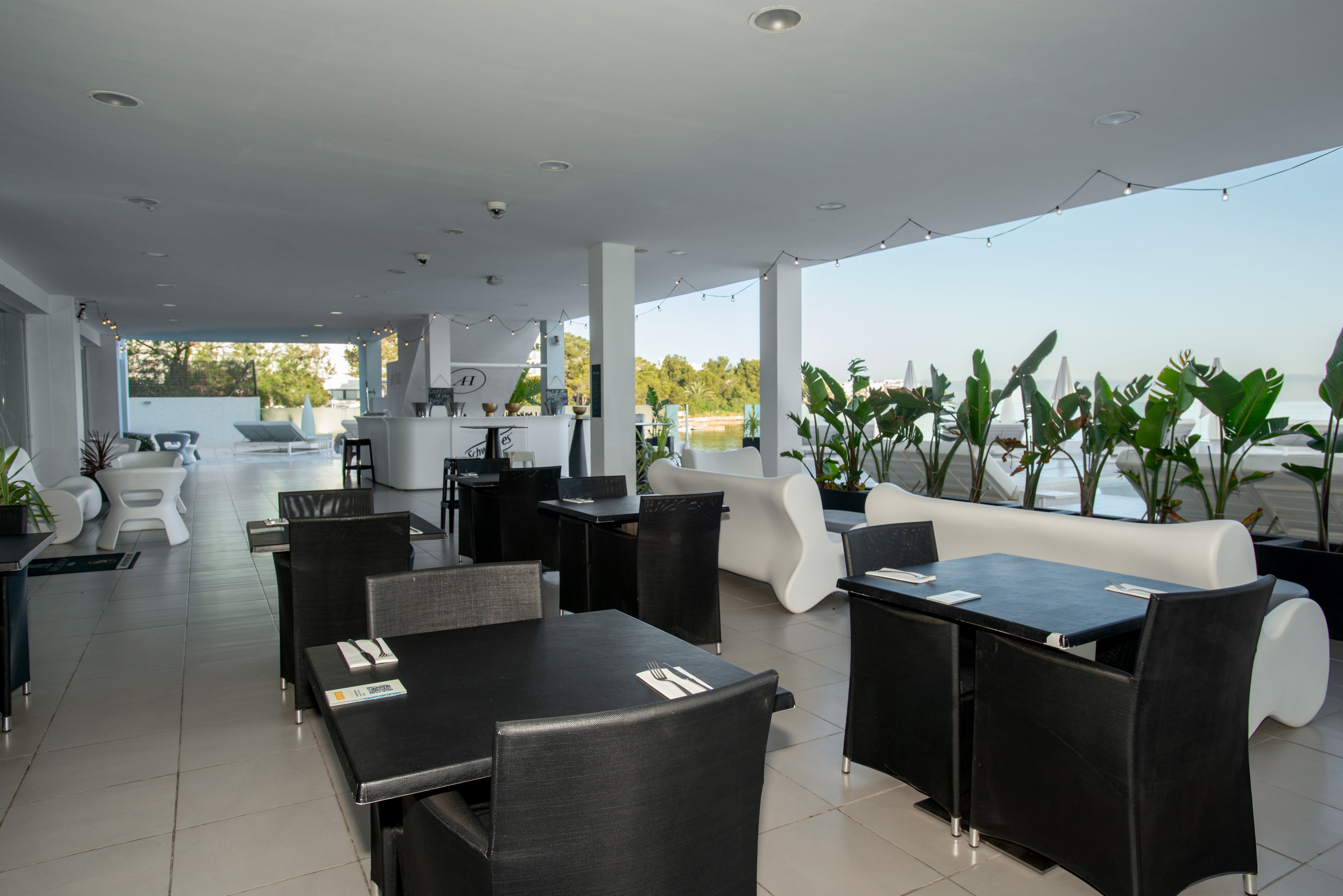 Contemporary black and white dining tables are arranged on an open-air restaurant terrace. The space overlooks a swimming pool and distant greenery under a clear sky. Sleek minimalist furniture and potted tropical plants create a stylish, resort-like atmosphere. The setting appears calm and ready for guests, with place settings neatly arranged on the tables.