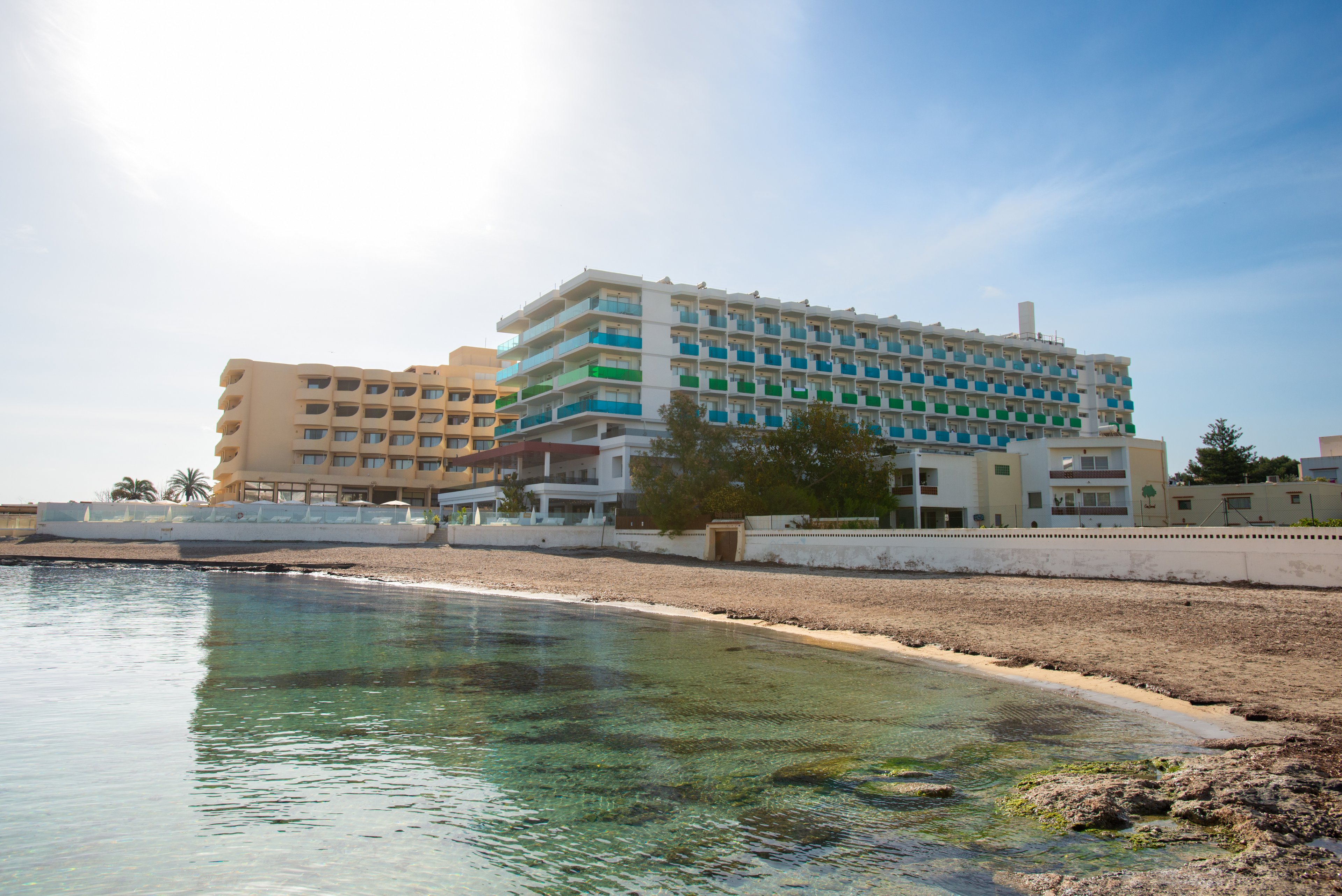 A large modern hotel complex stands along a quiet sandy beach beside clear, shallow water. The multi-story building features rows of balconies facing the sea, suggesting rooms with ocean views. Sunlight creates a bright, warm atmosphere with a clear blue sky overhead. The shoreline includes patches of rocks and seaweed in the transparent water, emphasizing the coastal setting.