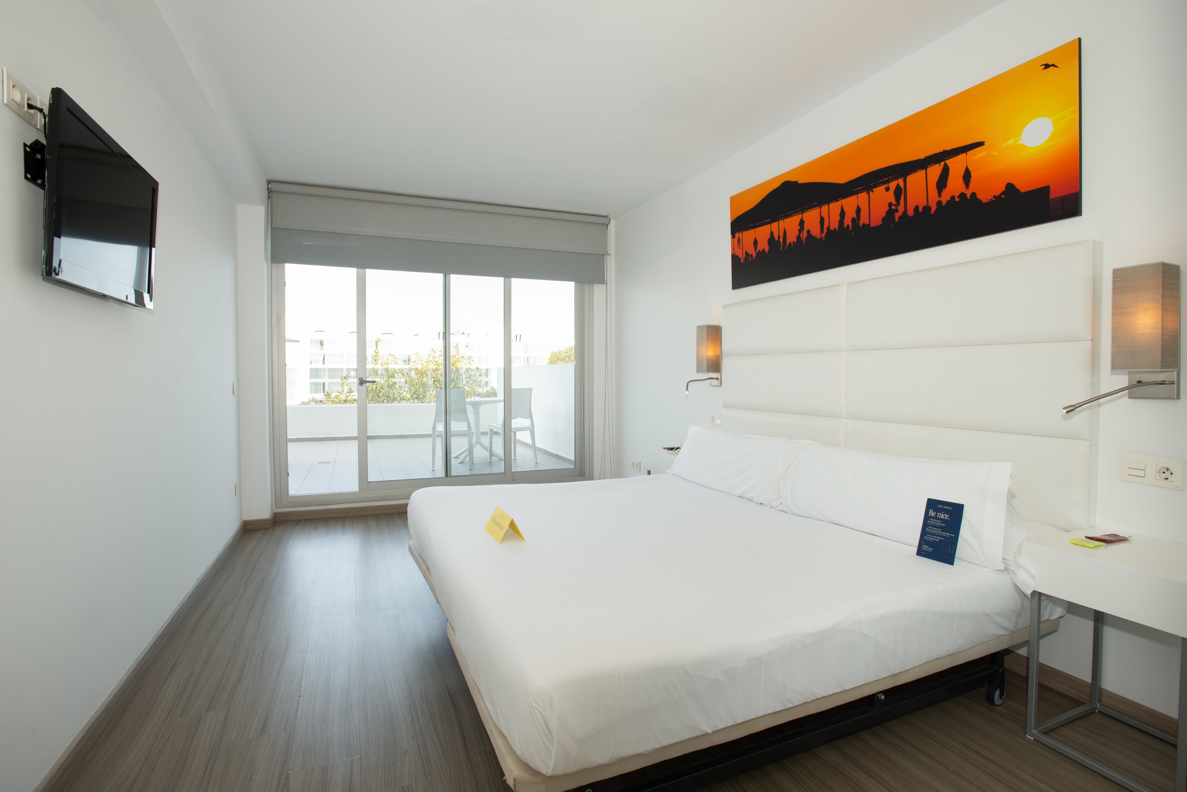 Bright, contemporary hotel room featuring a large white bed and minimalist furnishings. A wide glass sliding door opens onto a private balcony with outdoor chairs and city views. Warm wall lamps and a colorful sunset artwork above the headboard add contrast to the otherwise neutral decor. A flat-screen TV is mounted on the wall, and the room appears clean, spacious, and inviting.
