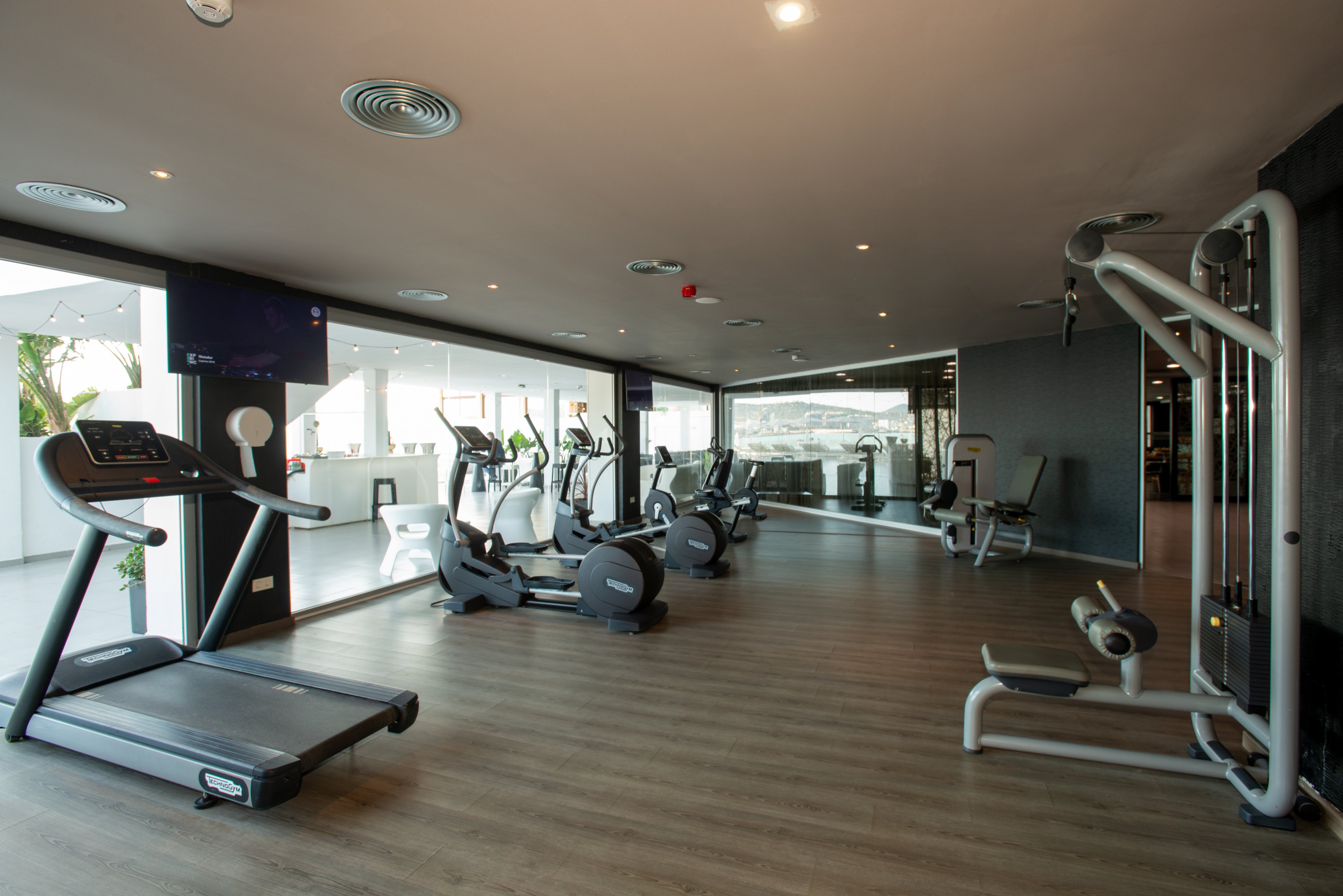 A spacious, modern gym interior features several treadmills, stationary bikes, and elliptical machines arranged along large windows. The room has wood-style flooring, recessed ceiling lights, and neutral-colored walls that create a clean, contemporary look. Strength-training machines are positioned on the right side, adding variety to the workout options. The gym appears empty and well-organized, emphasizing an inviting and professional fitness environment.
