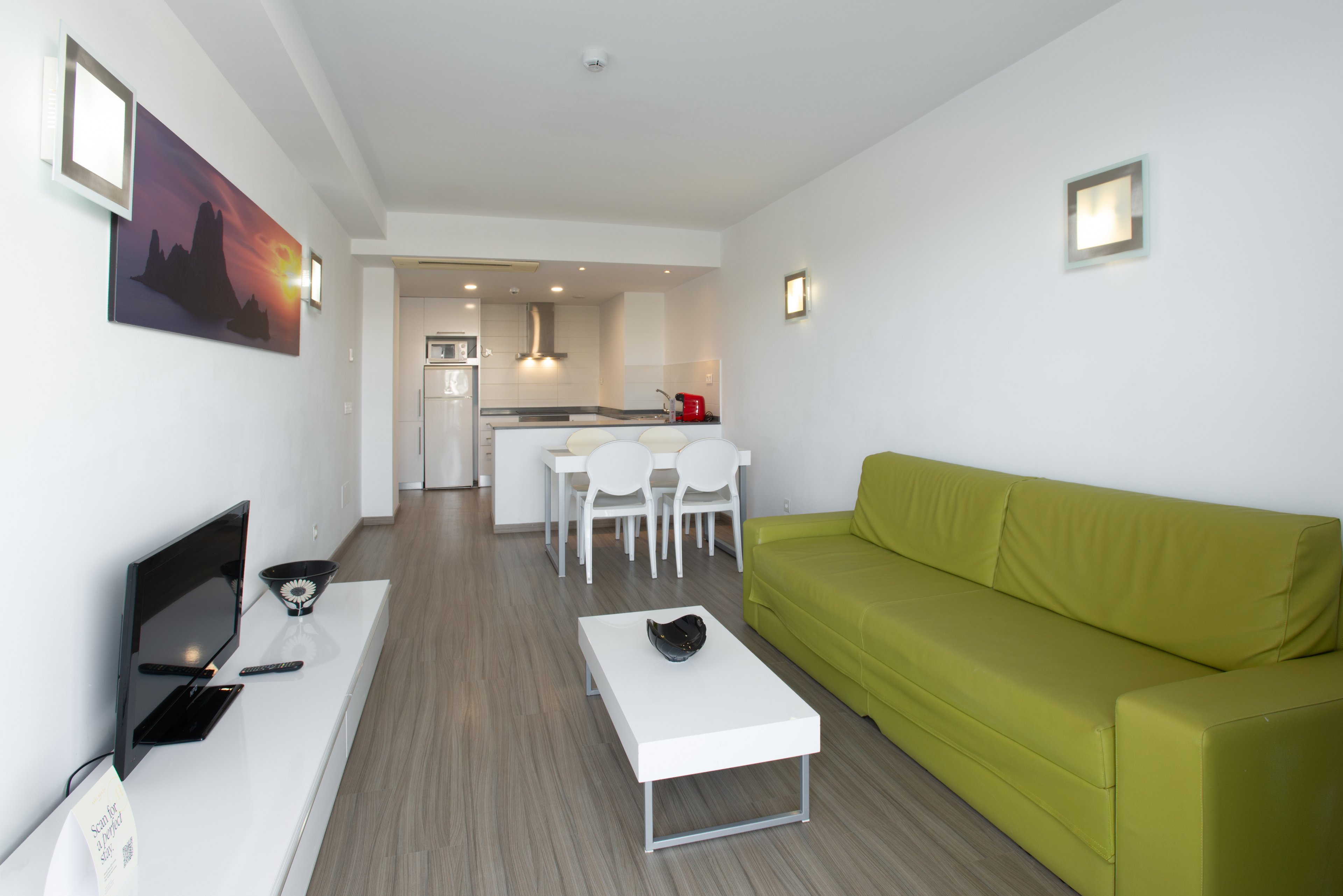 Bright contemporary living room with a green sofa faces a low white TV stand and flat-screen television. The open-plan space extends into a compact kitchen and dining area with white chairs and table. Minimalist decor, clean white walls, and wood-look flooring create a fresh, modern atmosphere. Recessed wall lights and a colorful landscape artwork add subtle visual interest to the interior.