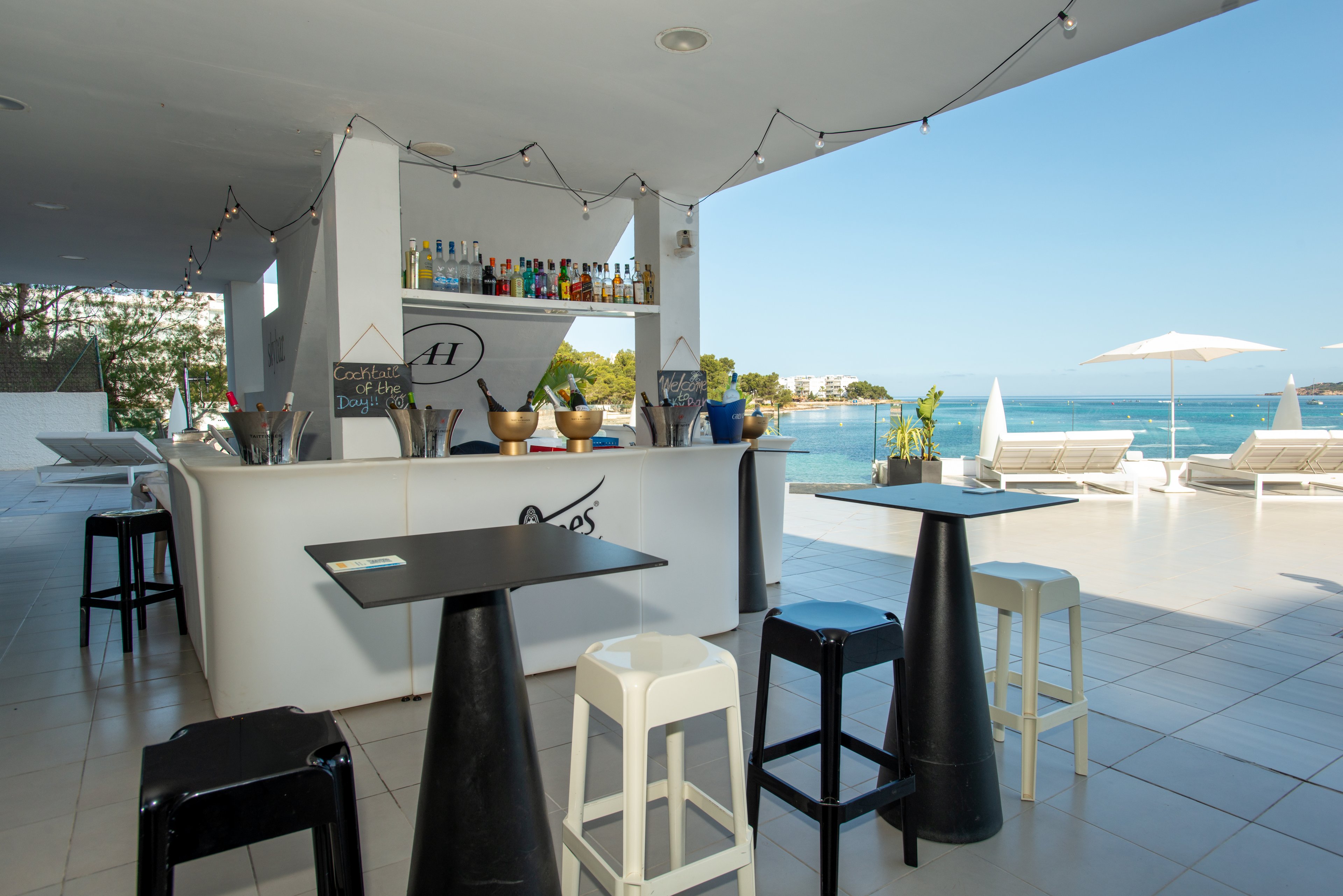 A contemporary open-air bar stands on a tiled terrace overlooking a calm blue sea. High stools and small tables are arranged around the minimalist white counter, which is stocked with colorful bottles and bar tools. String lights hang from the ceiling, adding a festive touch to the clean, modern design. In the background, white sun loungers and umbrellas line the waterfront, creating a relaxed vacation atmosphere.