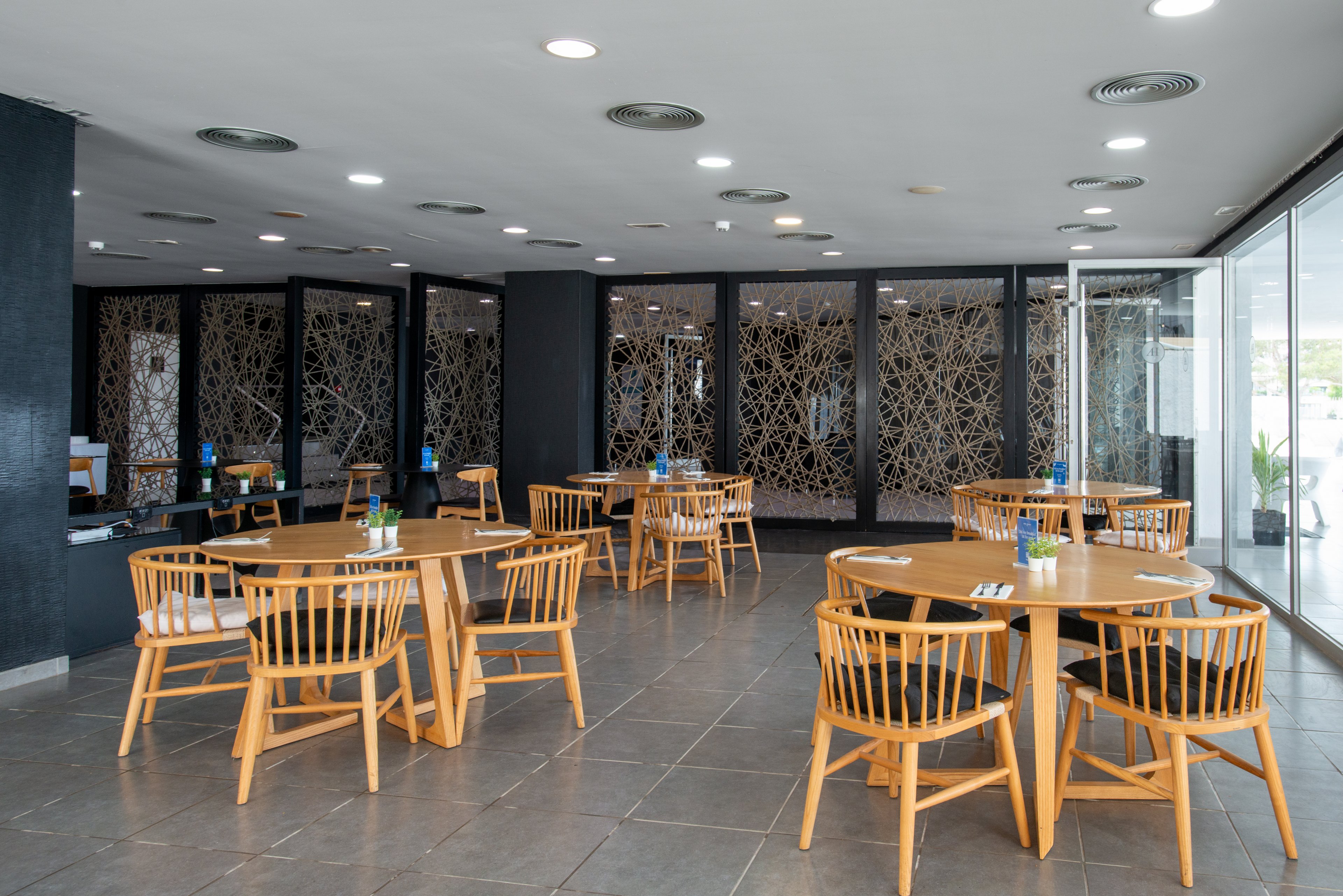 A contemporary restaurant dining area features several round wooden tables with matching wooden chairs. The space is empty of people, emphasizing the clean and orderly arrangement of the furniture. Dark walls and decorative partition panels contrast with the light wood tones and gray tiled floor. Ceiling spotlights and large windows provide bright, even lighting throughout the room.