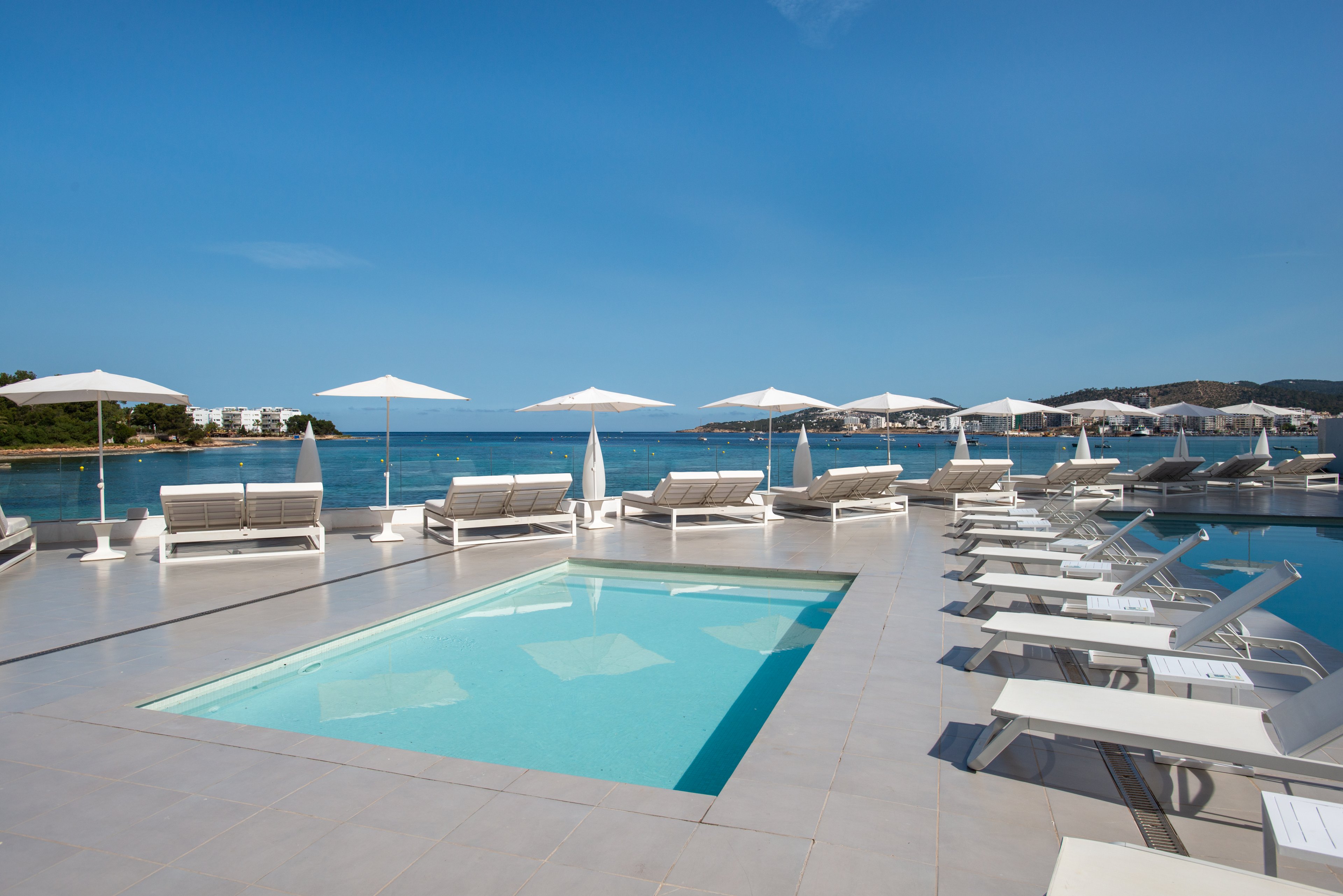 A sleek, modern pool deck overlooks a calm seaside bay under a clear blue sky. White sun loungers and umbrellas line the edge of an infinity-style pool, creating a minimalist and luxurious atmosphere. The light-colored tiles and turquoise water contrast with the deep blue ocean in the background. No people are present, emphasizing tranquility and relaxation in this coastal resort setting.