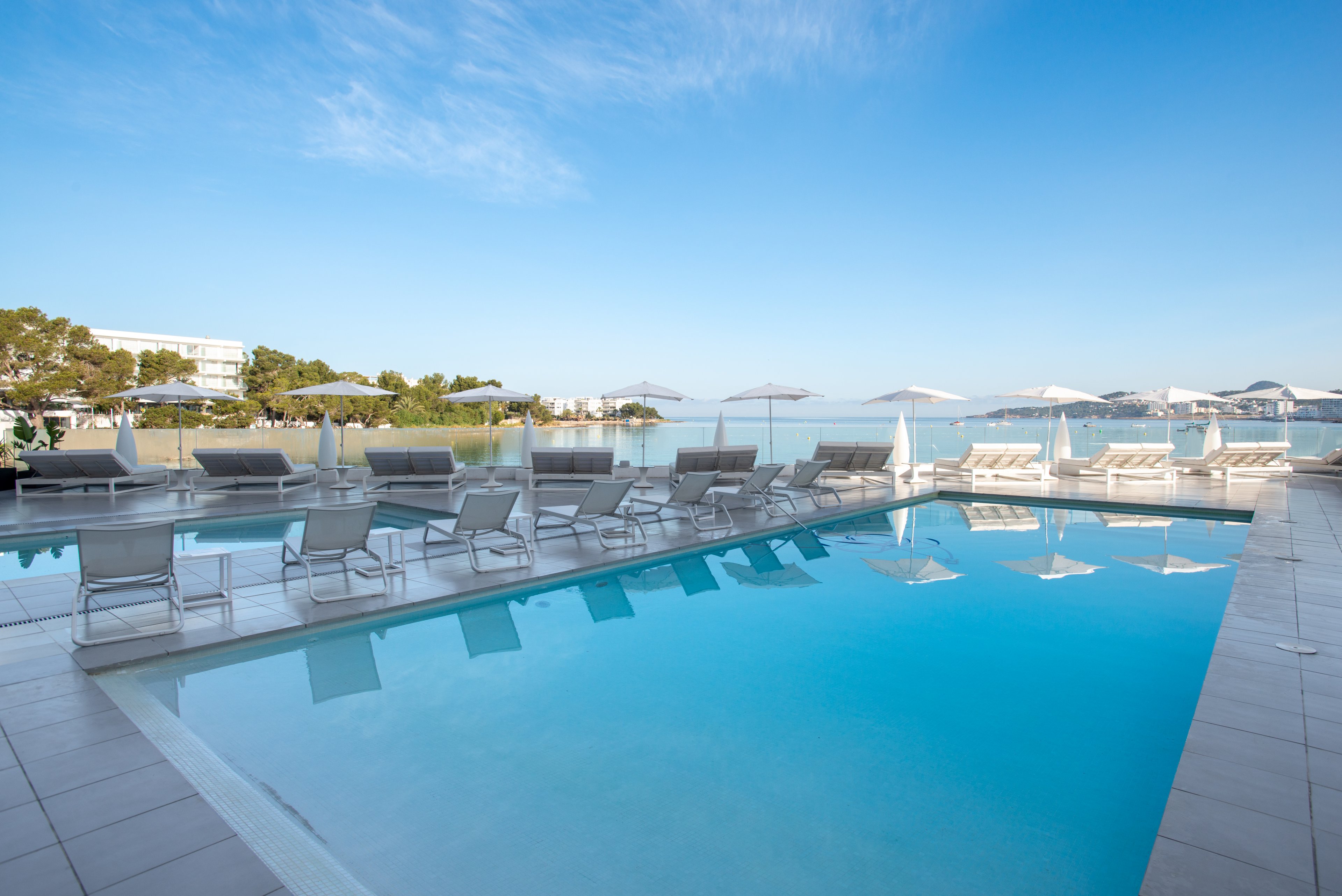 A serene outdoor swimming pool overlooks a calm seaside bay under a clear blue sky. White sun loungers and umbrellas line the pool deck, reflecting in the still turquoise water. The minimalist, modern design and light color palette create a luxurious resort atmosphere. Distant buildings and greenery frame the horizon, enhancing the sense of a peaceful coastal retreat.