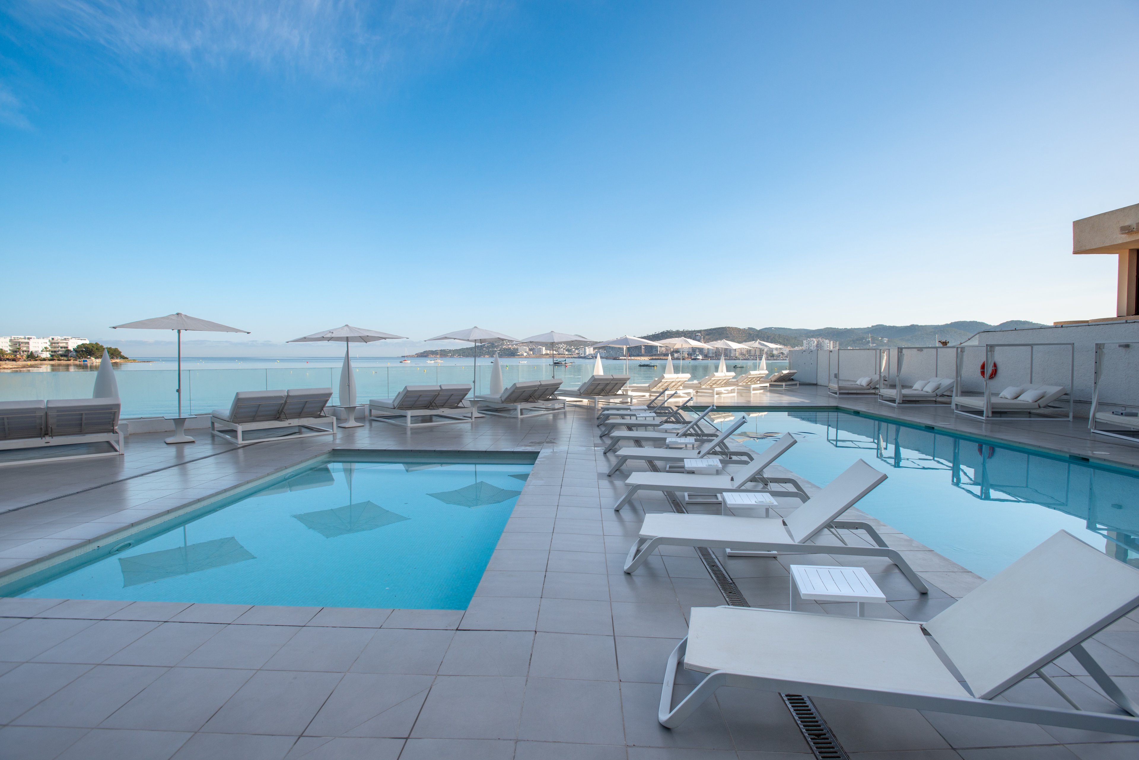 A sleek rooftop pool area features an infinity-edge pool and a smaller adjacent pool surrounded by white lounge chairs. The scene overlooks a calm coastal bay with distant buildings and hills under a clear blue sky. White umbrellas line the deck, and glass railings provide unobstructed views of the water. The overall atmosphere is tranquil and minimalist, suggesting a luxurious resort or hotel setting.
