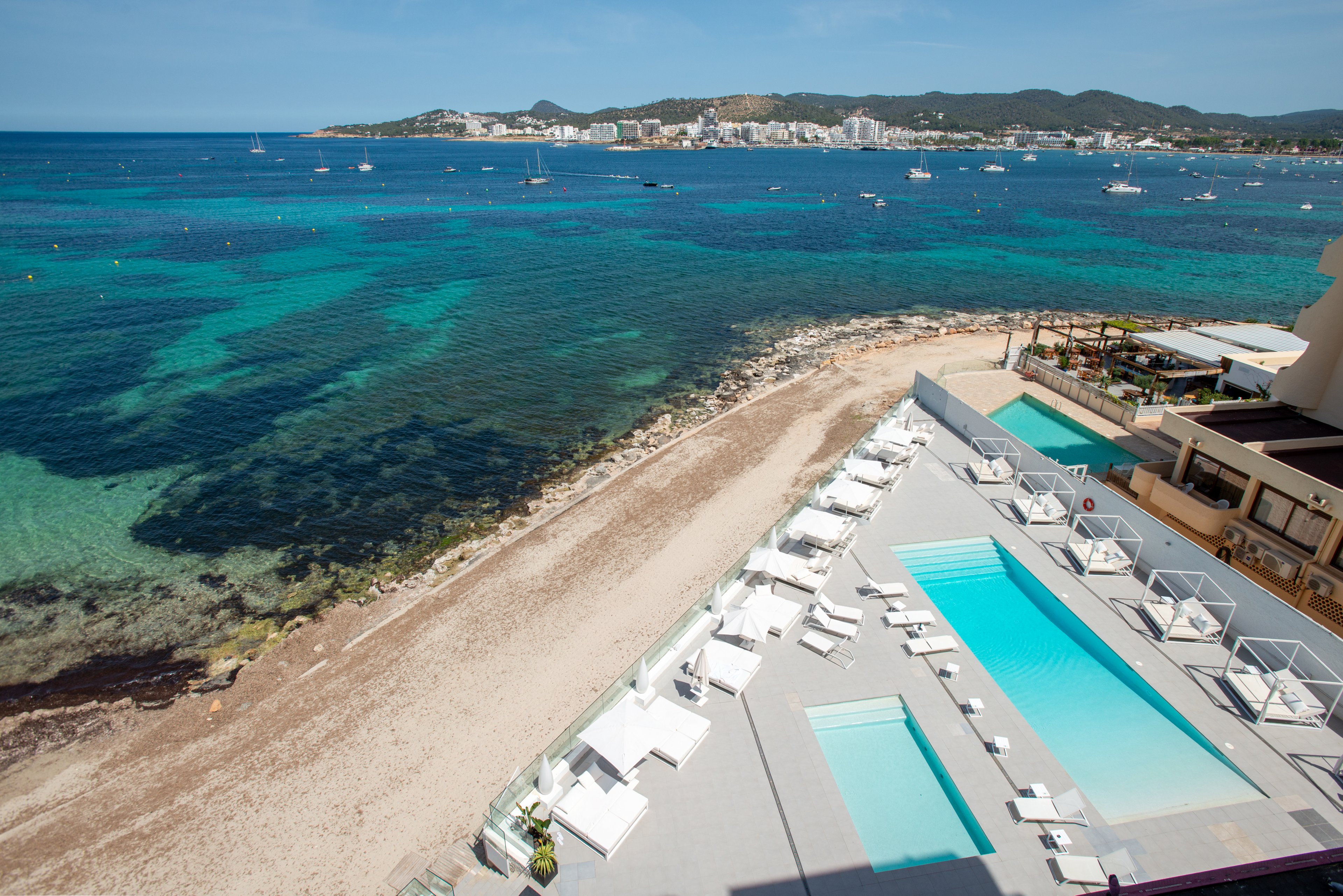 A contemporary beachfront resort features a long rectangular swimming pool and sun loungers arranged neatly on a light-colored terrace. The scene overlooks a clear turquoise sea with gentle waves and a sandy shoreline in the foreground. In the distance, boats dot the water and low-rise buildings line the coastline under a bright blue sky. The overall atmosphere is calm, sunny, and inviting, ideal for seaside relaxation.