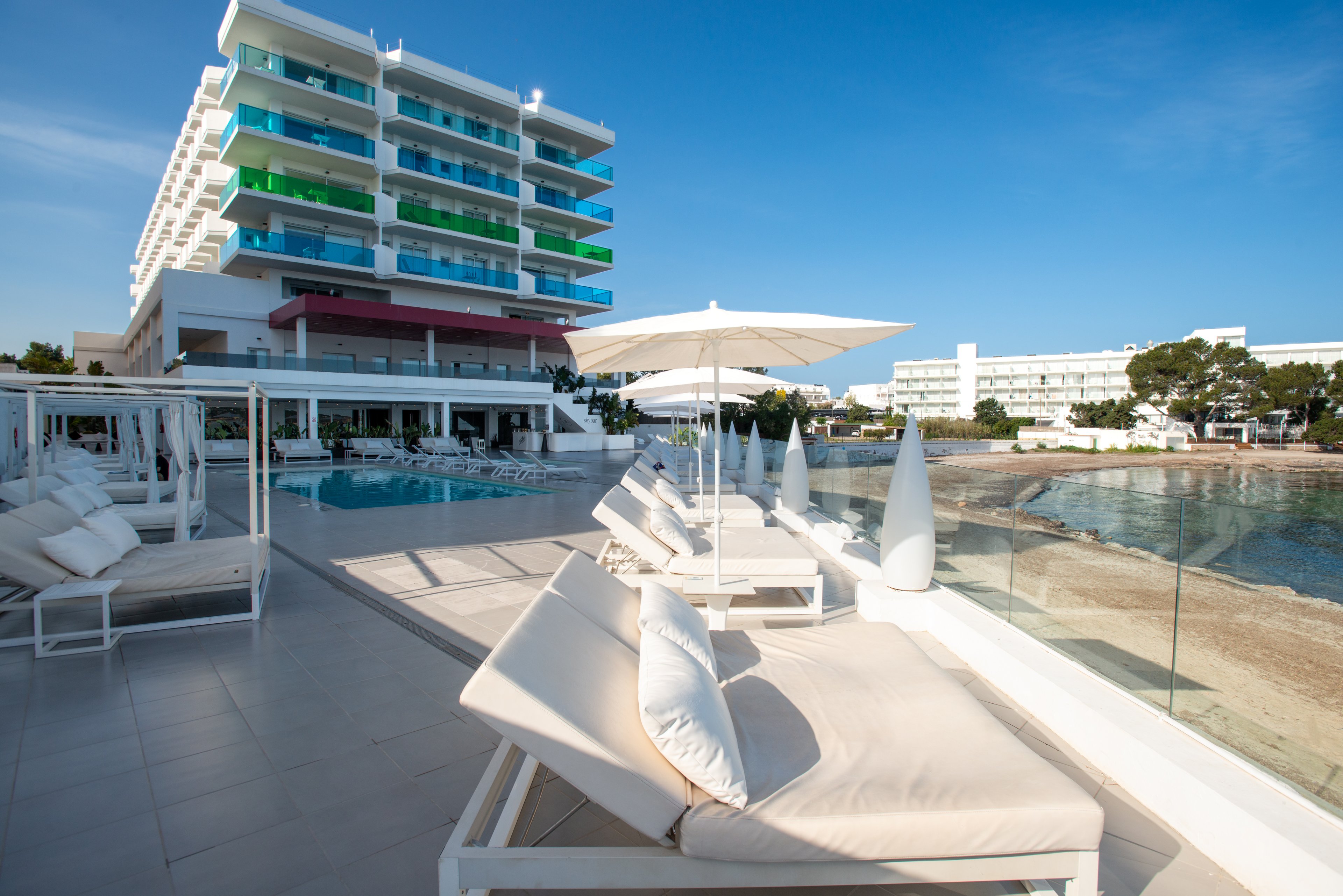 A contemporary multi-story hotel overlooks a calm beachfront with a spacious terrace in the foreground. White cushioned sun loungers and umbrellas line the tiled deck beside a clear swimming pool. The scene is bathed in bright daylight under a clear blue sky, creating a relaxed vacation atmosphere. Nearby buildings and trees frame the coastal setting along the water’s edge.