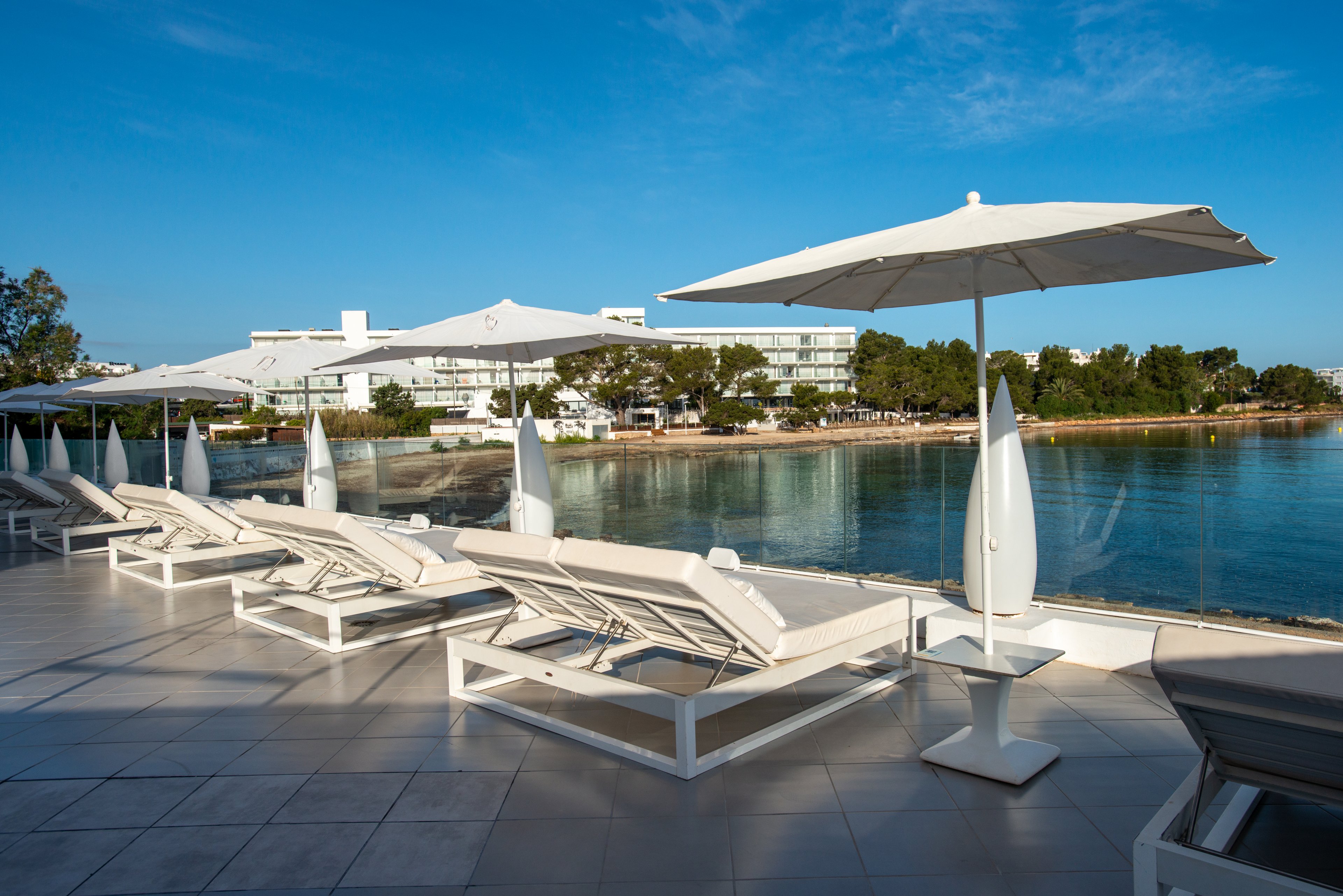 A row of white sun loungers and umbrellas lines a tiled terrace overlooking a calm waterfront. The scene appears to be part of a modern seaside resort with low-rise buildings in the background. Clear blue sky and still water create a peaceful, inviting atmosphere. The area is empty of people, emphasizing relaxation and tranquility.