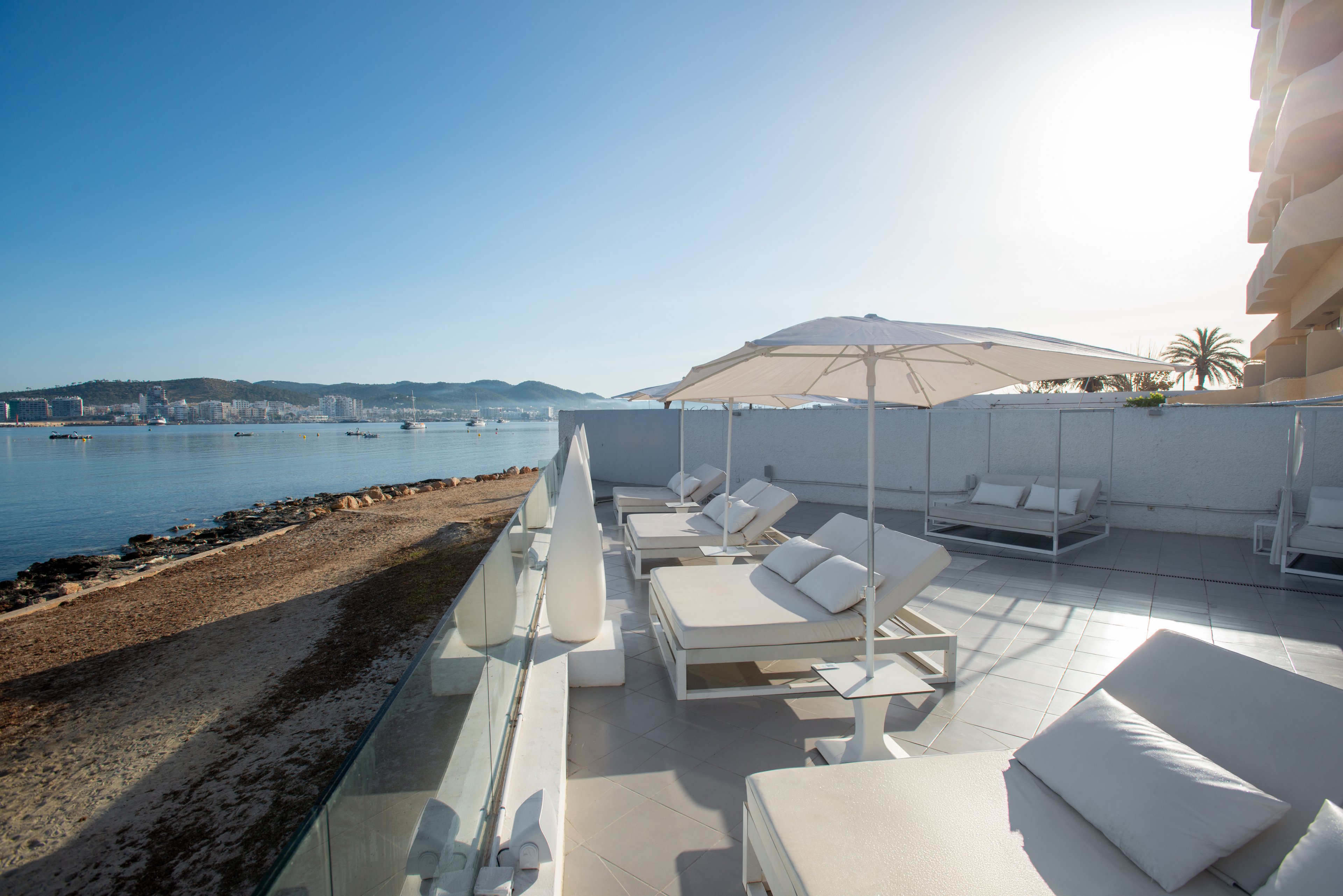 A spacious hotel terrace overlooks a calm seaside bay under a clear blue sky. White sun loungers and umbrellas are neatly arranged along the tiled deck, facing the water. Glass railings separate the terrace from the sandy shoreline and rocky edge below. Distant buildings and boats are visible across the water, enhancing the relaxed coastal resort atmosphere.