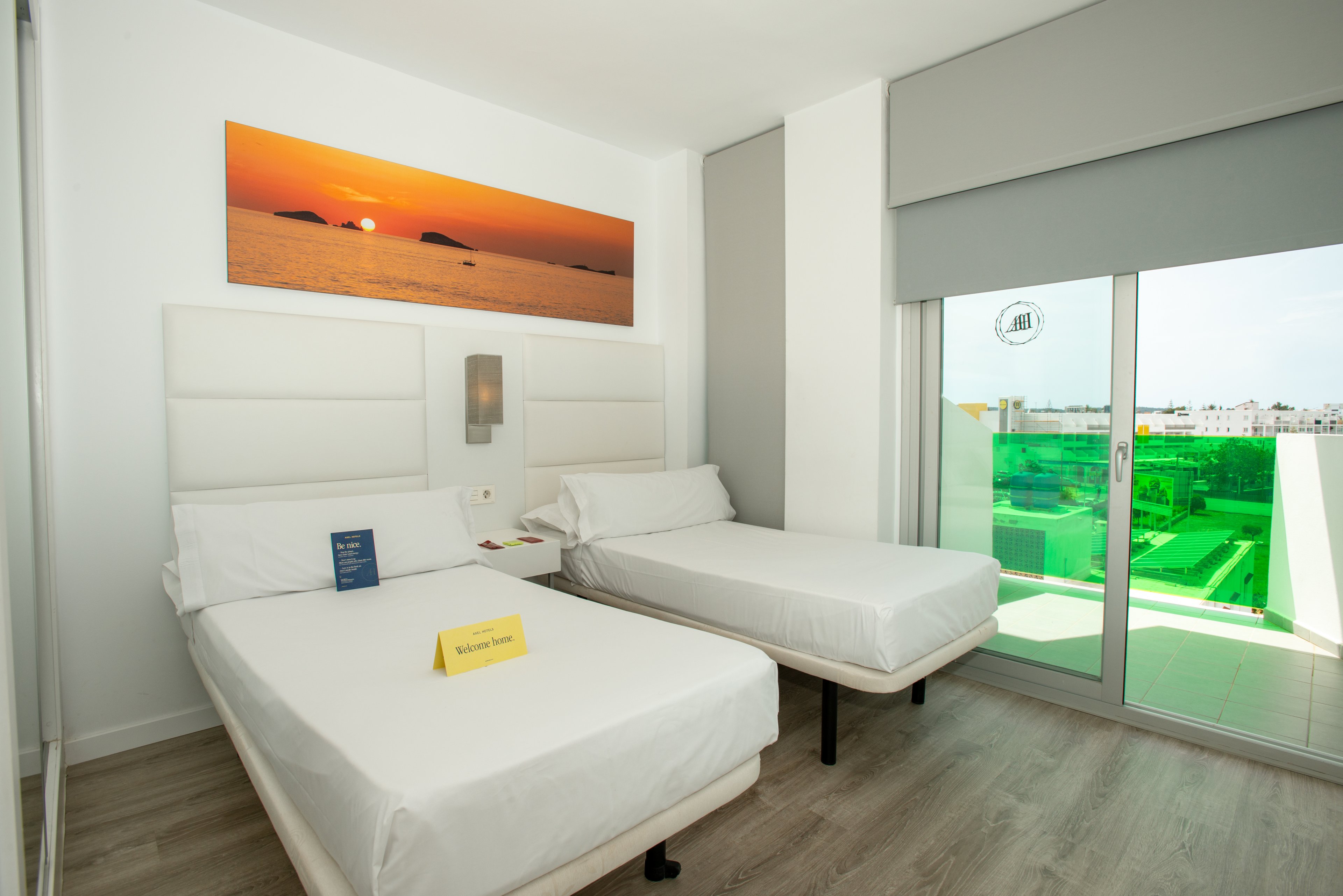 Bright contemporary hotel room featuring two single beds with white bedding and padded headboards. A large panoramic sunset photograph decorates the wall above one bed, adding warm color to the minimalist interior. Floor-to-ceiling glass doors open onto a balcony with a city and rooftop view, filtered by green-tinted panels. The room has light wood flooring and neutral walls, creating a clean and modern atmosphere.