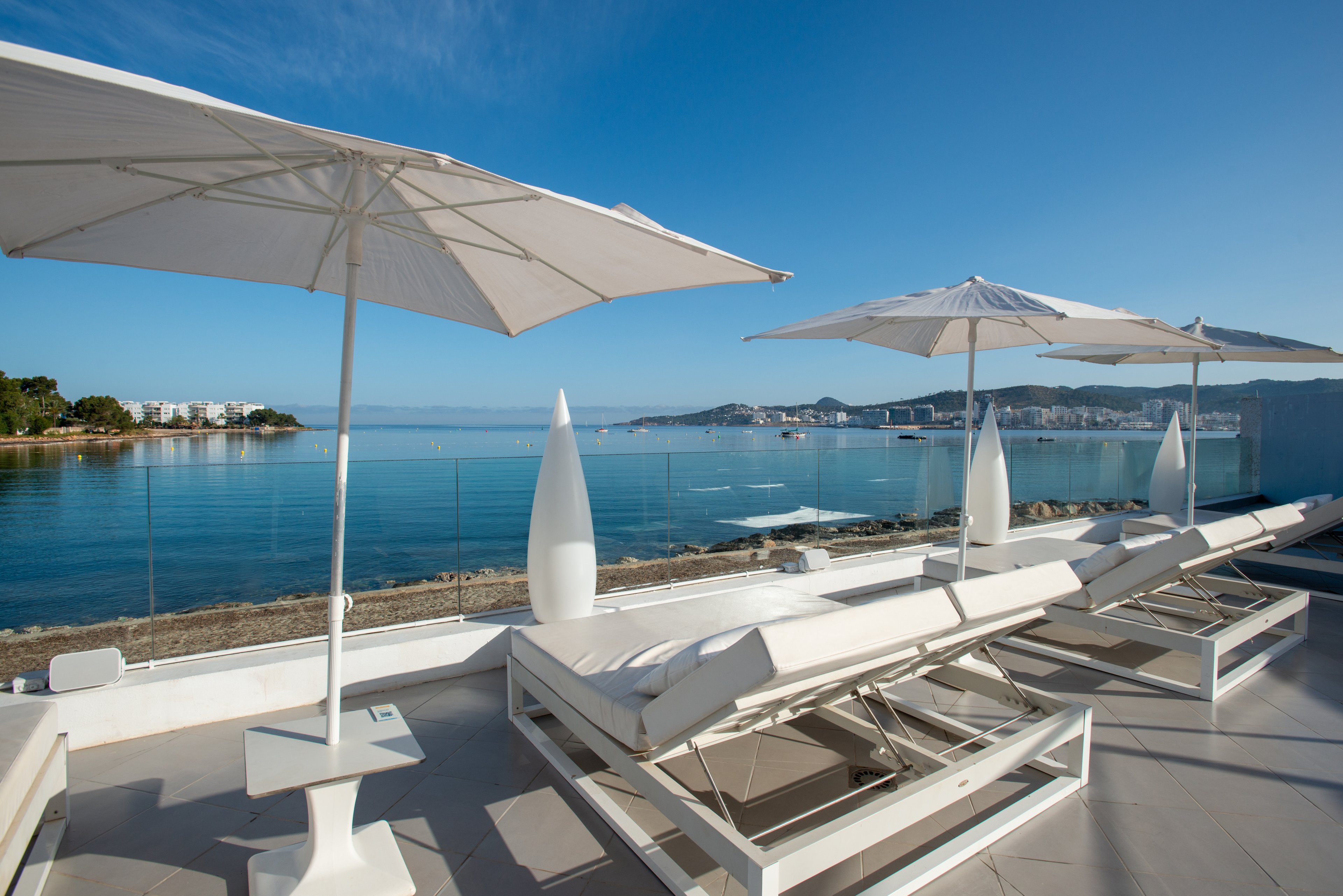 A contemporary rooftop terrace overlooks a calm blue sea under a clear sky. White cushioned sun loungers are arranged in a row beside sleek side tables and tall sculptural lights. Large white umbrellas provide shade and emphasize the minimalist, luxurious design. In the distance, low-rise coastal buildings line the shoreline, enhancing the relaxed vacation atmosphere.