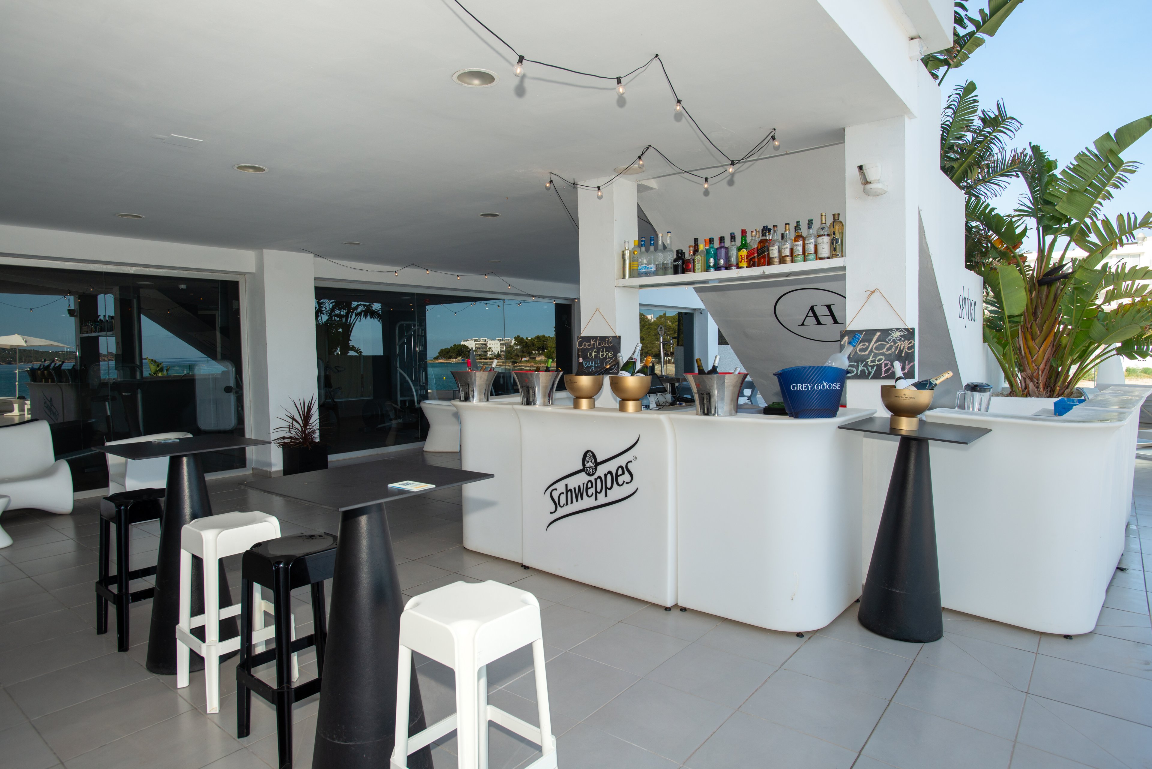 An open-air bar area features a sleek white counter prominently displaying the Schweppes name. Various liquor bottles are arranged on shelves behind the bar, with chalkboard signs and cocktail shakers adding to the mixology theme. Modern stools and cone-shaped tables provide seating in the bright, minimalist space. Large windows and tropical plants suggest a relaxed, resort-like atmosphere by the water.