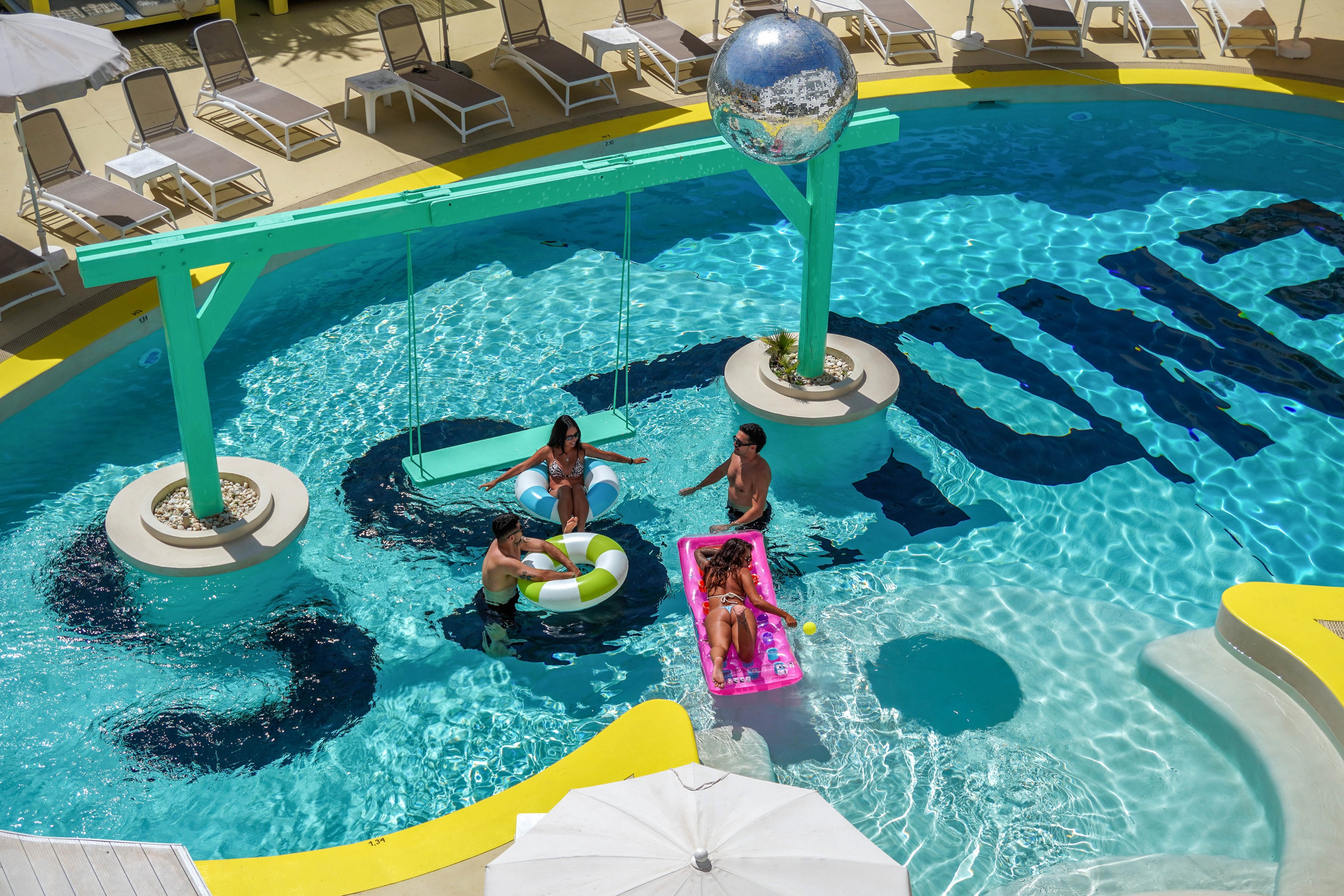 A group of young adults relaxes together in a bright turquoise resort swimming pool. Two people float on inflatable pool loungers while another stands in the water and one sits on a swing suspended over the pool. Lounge chairs and umbrellas surround the pool deck, suggesting a sunny vacation setting. The overall scene feels playful and summery with bold colors and a festive atmosphere.