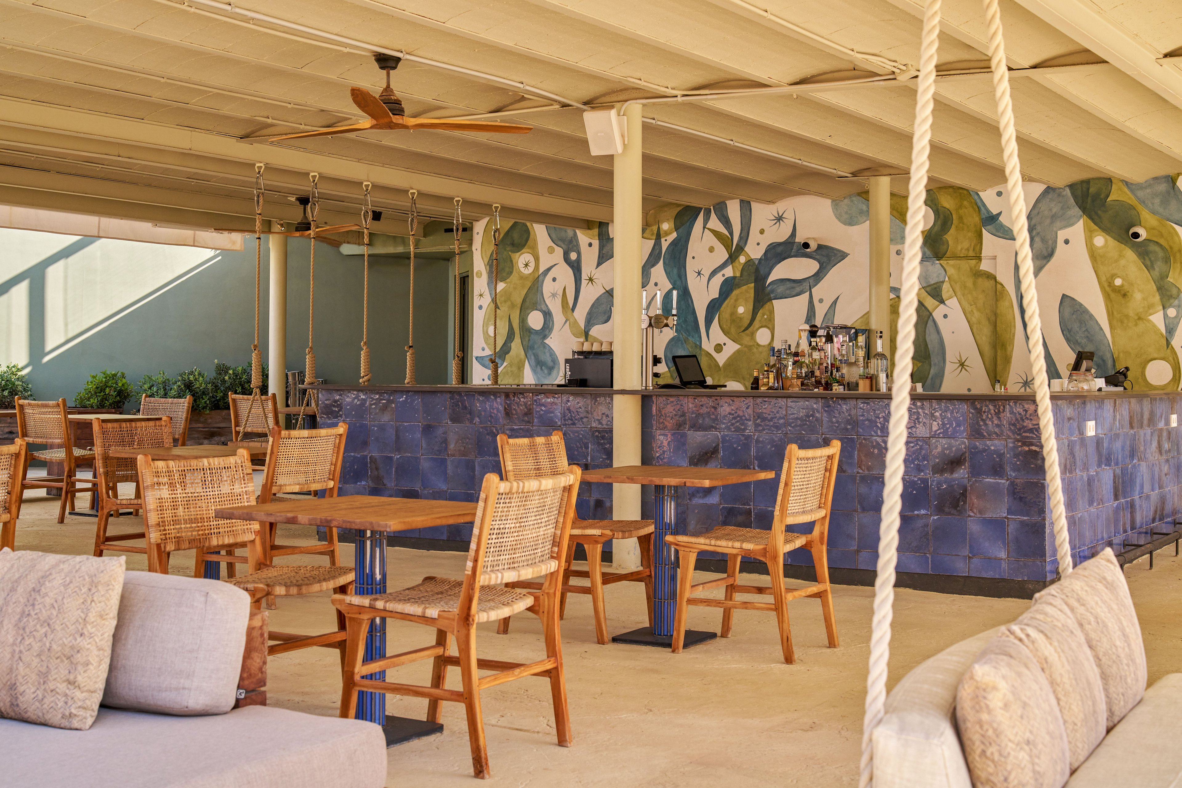 An open-air bar area features wooden tables and wicker chairs arranged on a smooth floor. A tiled bar counter with a colorful abstract mural behind it forms the focal point of the space. Ceiling fans and rope swings add a relaxed, coastal atmosphere. Soft neutral tones and natural materials create a warm, inviting setting for guests.
