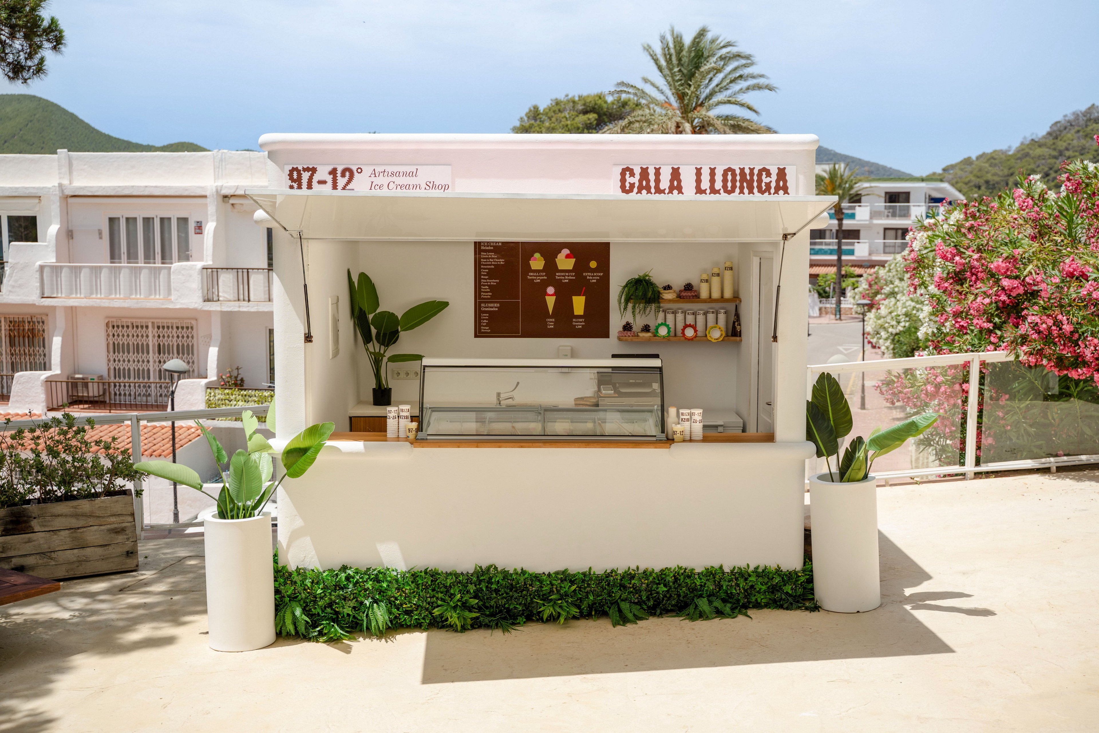A small white ice cream kiosk labeled Cala Llonga stands in a sunny outdoor setting. The stand features a central freezer display, a menu board with illustrated cones and cups, and neatly arranged jars and toppings. Potted green plants frame the kiosk, while residential buildings, palm trees, and flowering bushes appear in the background. The scene suggests a relaxed, coastal holiday atmosphere.
