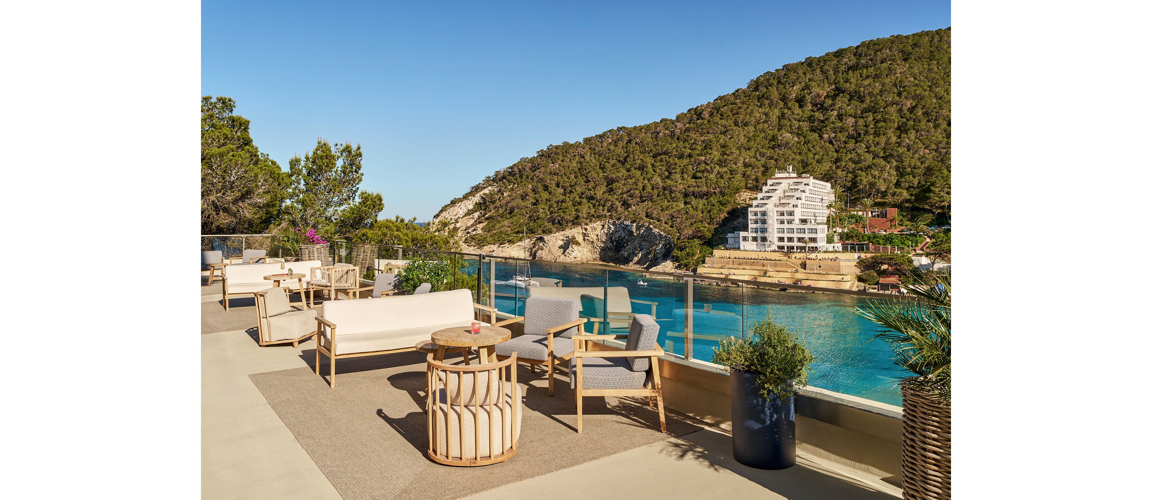 An elegant outdoor terrace with modern lounge furniture overlooks a calm turquoise bay. Glass railings frame the view of forested hills and white seaside buildings across the water. Potted plants and neutral-toned seating create a relaxed, contemporary atmosphere under a clear blue sky. The scene suggests a peaceful vacation or resort setting by the coast.