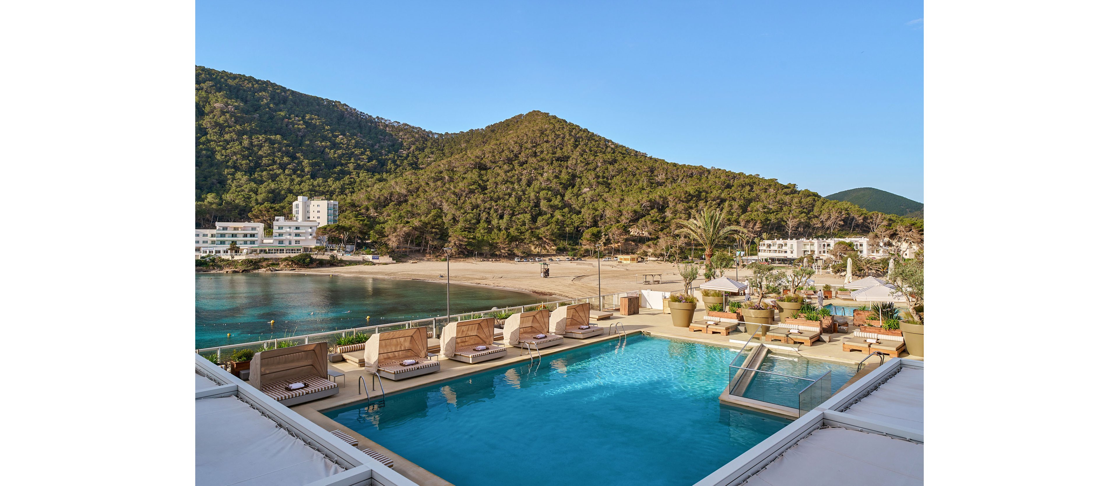 An elevated outdoor swimming pool overlooks a calm coastal bay and sandy beach. Lounge chairs and cabanas surround the pool, creating a relaxing vacation atmosphere. In the background, forested hills rise behind waterfront buildings under a clear blue sky. The scene suggests a tranquil Mediterranean-style resort setting ideal for leisure and sunbathing.