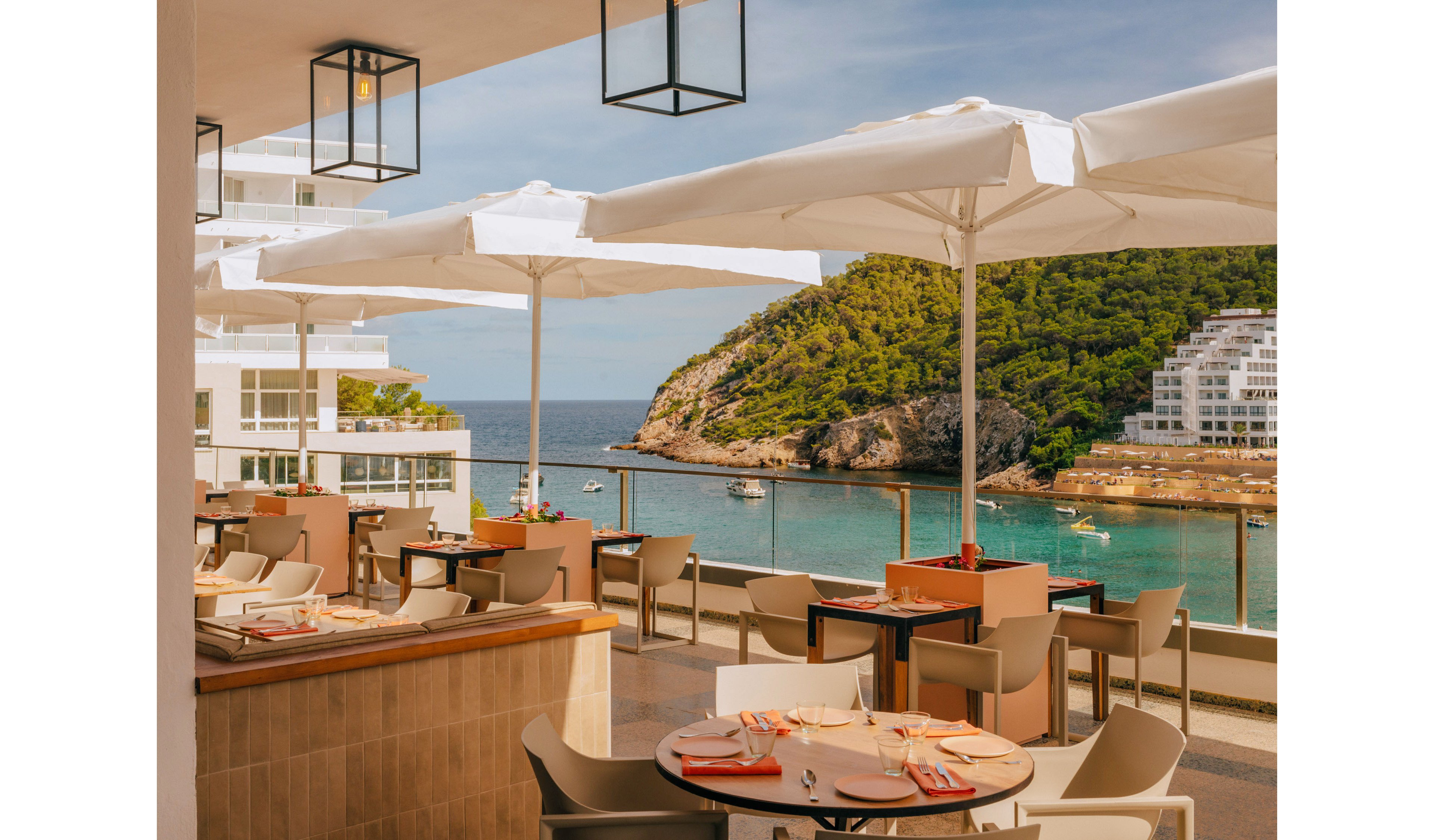 An open-air terrace restaurant features neatly set tables with plates, glasses, and orange napkins under large white umbrellas. The space is part of a modern waterfront hotel or resort, with clean architectural lines and balconies visible on the left. Beyond the terrace, a calm turquoise bay with small boats is framed by a lush green hillside and additional white buildings. Warm sunlight and soft colors create a relaxed, inviting vacation atmosphere.