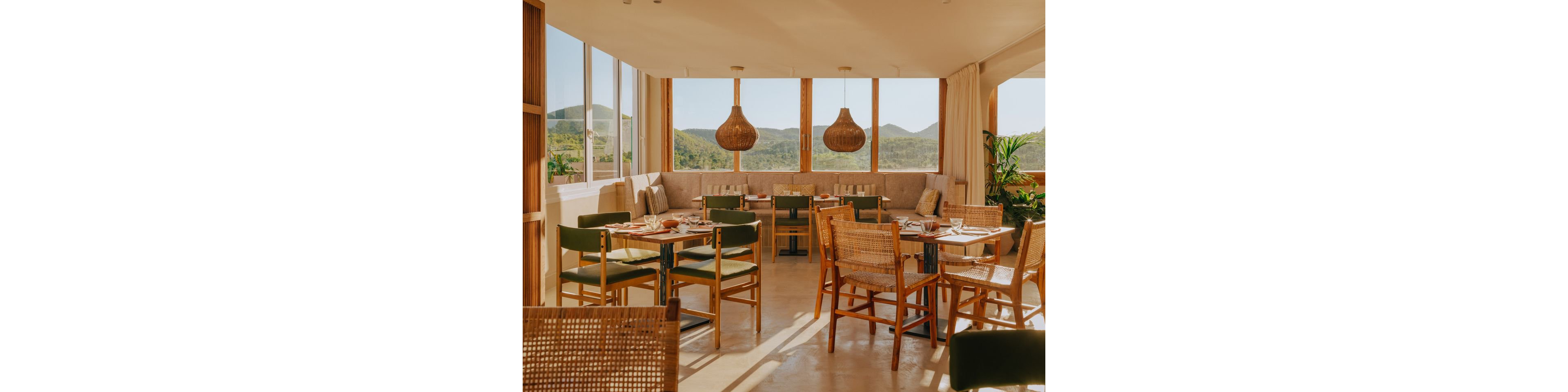 A bright, contemporary restaurant interior features wooden tables and mixed woven and upholstered chairs arranged neatly for dining. Large windows wrap around the space, revealing green hills and mountains in the distance. Warm natural light fills the room, highlighting neutral tones, rattan pendant lights, and soft bench seating along the wall. Potted plants add a touch of greenery, creating a relaxed and inviting atmosphere.
