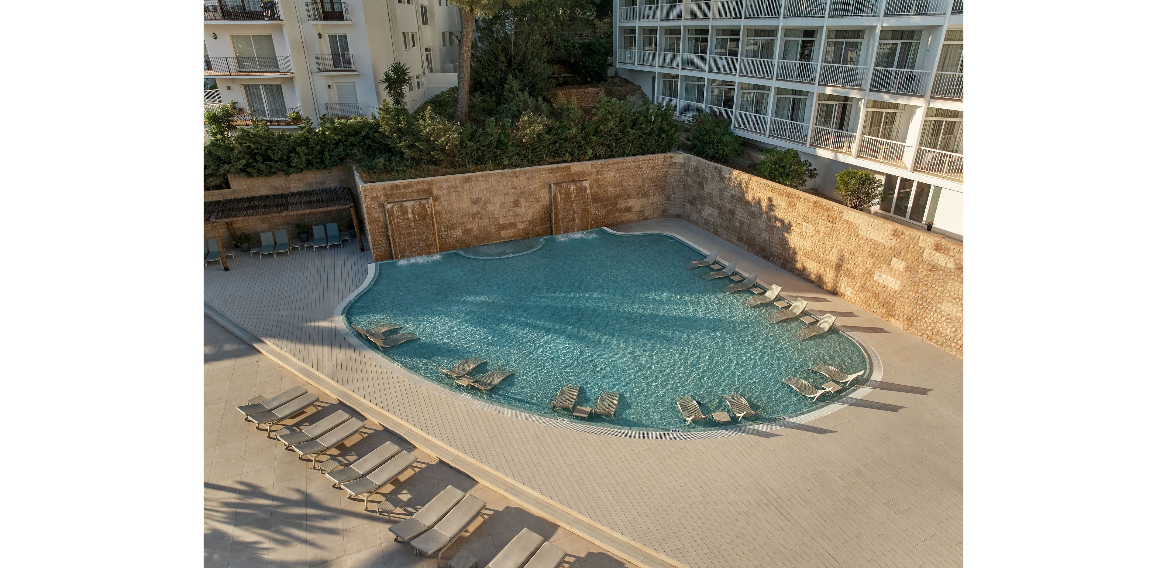 A curved outdoor swimming pool is surrounded by numerous sun loungers arranged neatly along the deck. The pool sits beside a multi-story hotel building with balconies overlooking the water. A tall stone wall and lush greenery provide privacy and a natural backdrop. The scene is calm and empty, suggesting an early morning or off-peak moment at the resort.